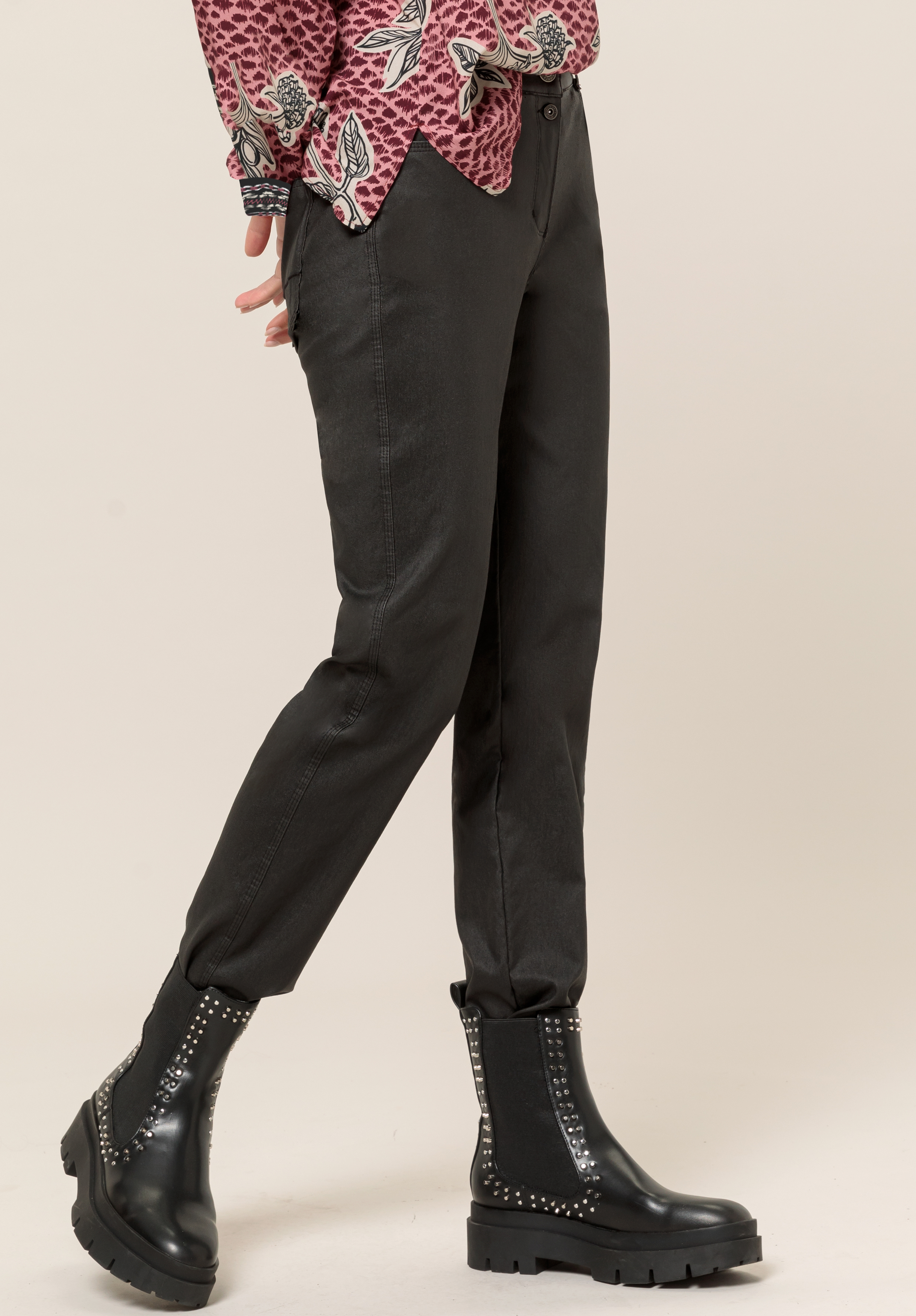 Eine Person in schwarzen Hosen, einem gemusterten Langarmhemd und schwarzen Stiefeletten mit silbernen Nieten steht vor einem schlichten hellen Hintergrund.