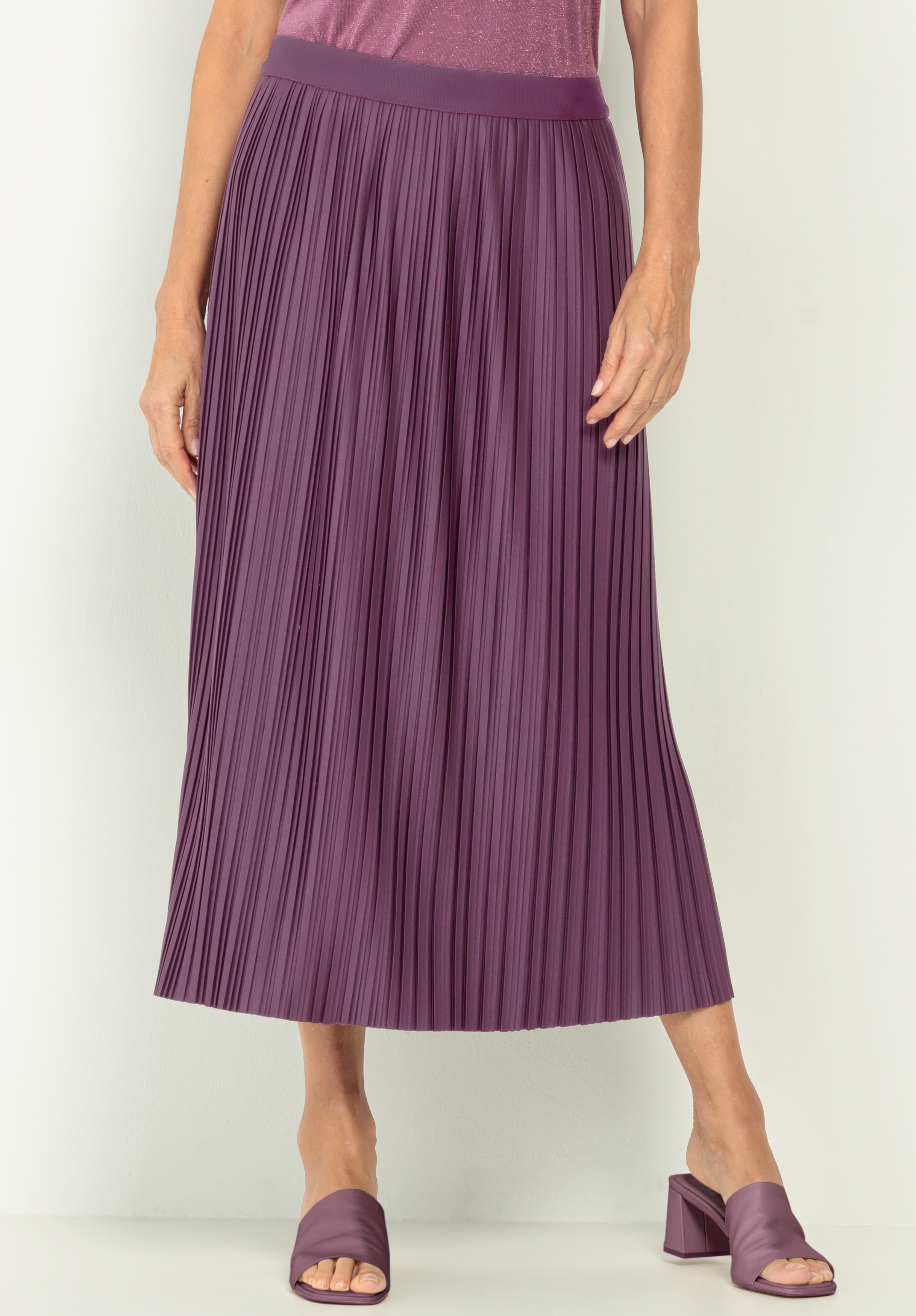 Eine Person, die einen gefalteten, knöchellangen lila Rock, passende lila offene Absätze und ein lila Oberteil trägt, steht vor einem schlichten hellen Hintergrund.
