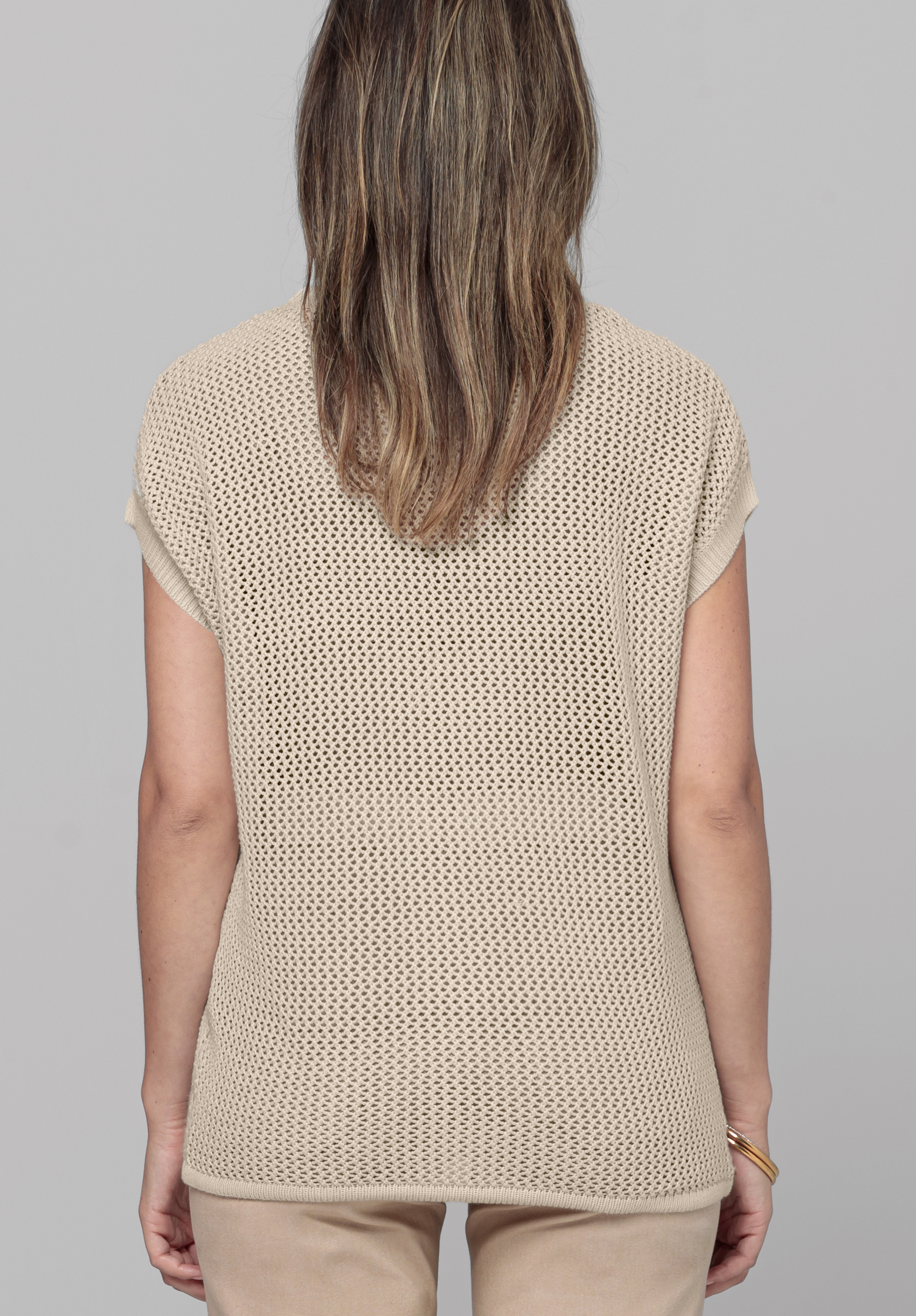 Eine Frau mit schulterlangem braunem Haar steht mit abgewandtem Gesicht da und trägt ein hellbeiges, kurzärmliges, perforiertes Strickoberteil und dazu passende beige Hosen vor einem schlichten grauen Hintergrund.
