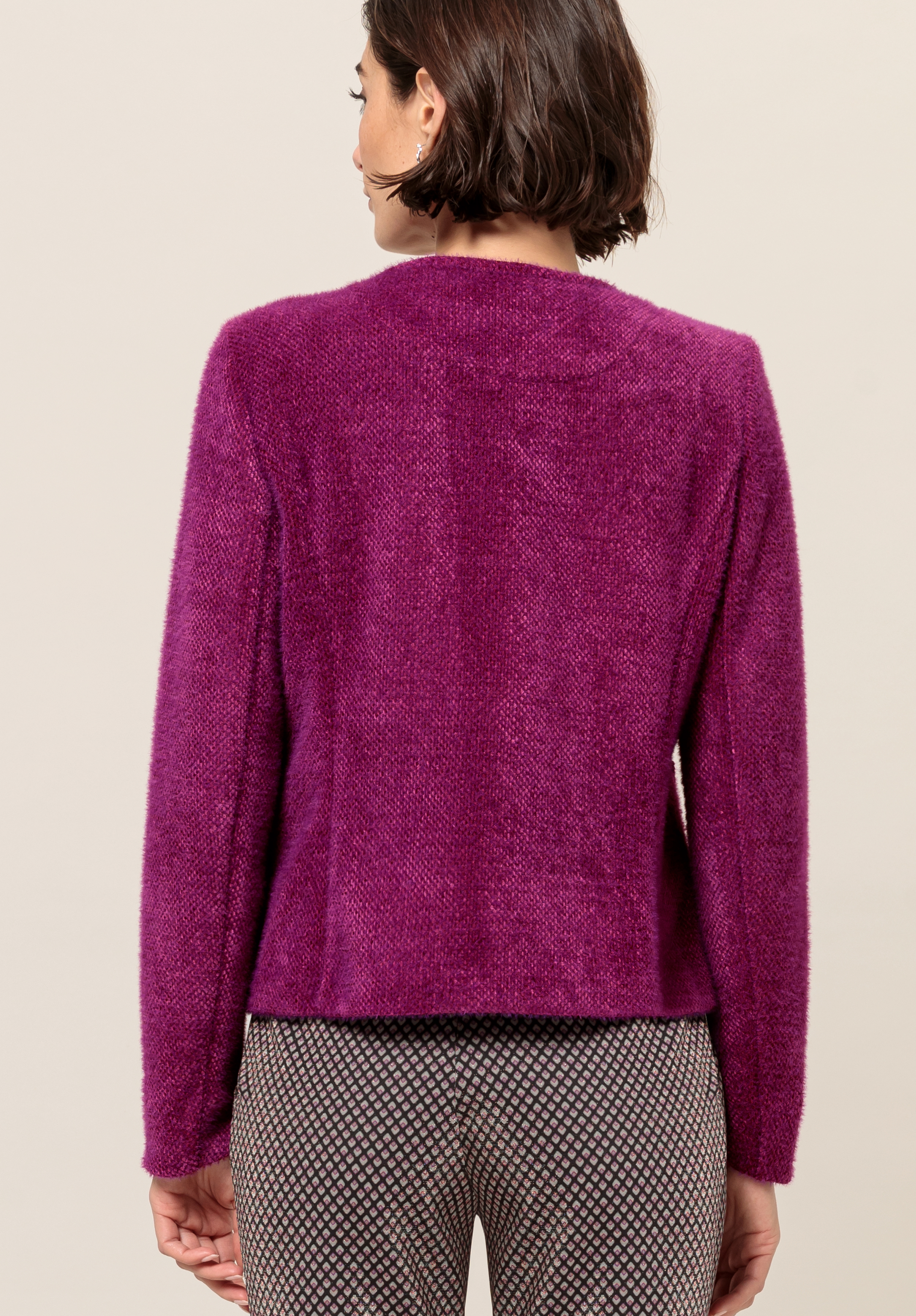 Von hinten ist eine Frau mit kurzen braunen Haaren zu sehen. Sie trägt einen strukturierten magentafarbenen Pullover und gemusterte graue Hosen und steht vor einem schlichten beigen Hintergrund.