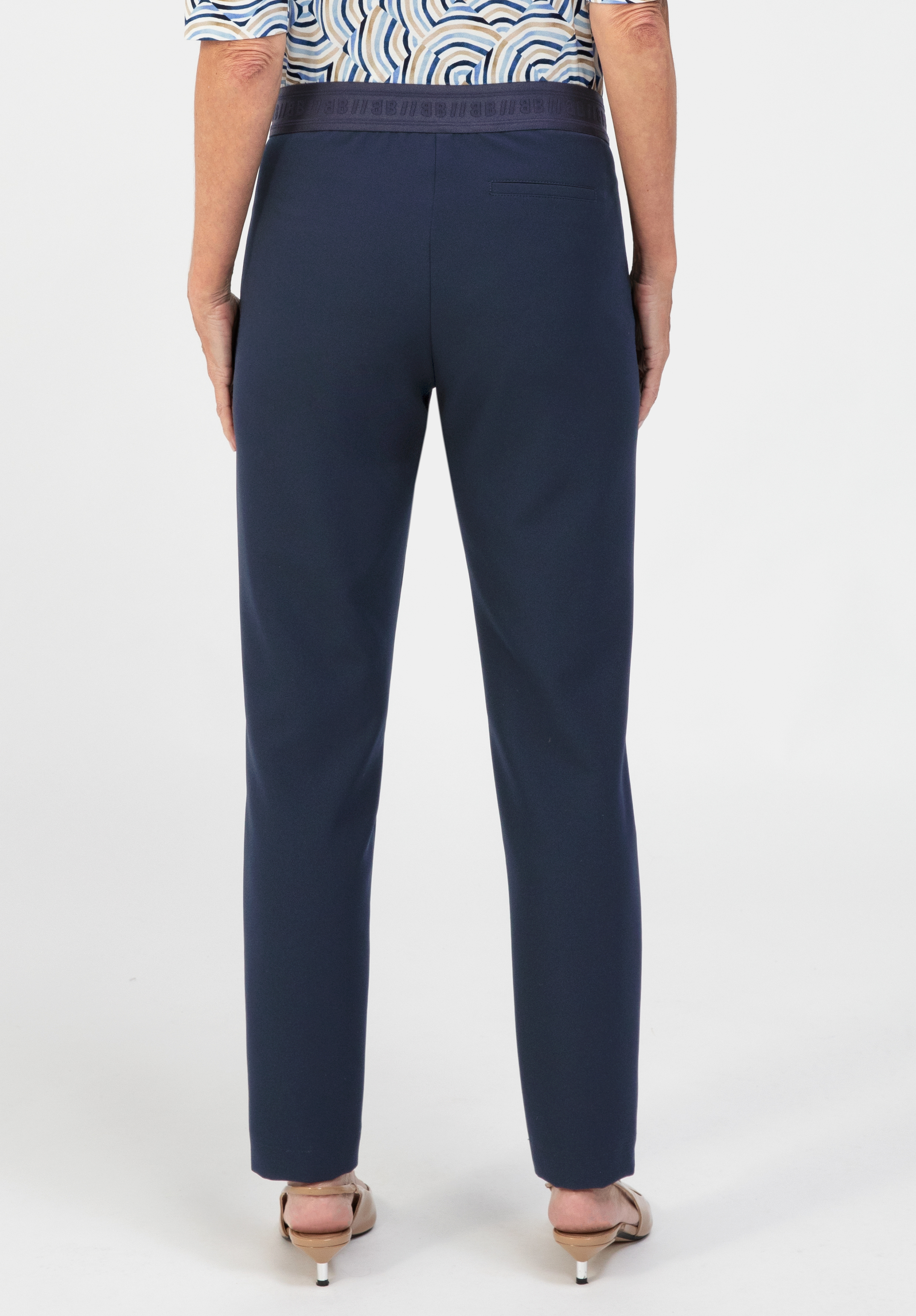 Eine Person ist von der Taille abwärts zu sehen; sie trägt eine marineblaue Hose mit geradem Bein und hellbraune Schuhe mit niedrigem Absatz und steht vor einem weißen Hintergrund. Der obere Teil eines gemusterten Hemdes ist teilweise zu sehen.