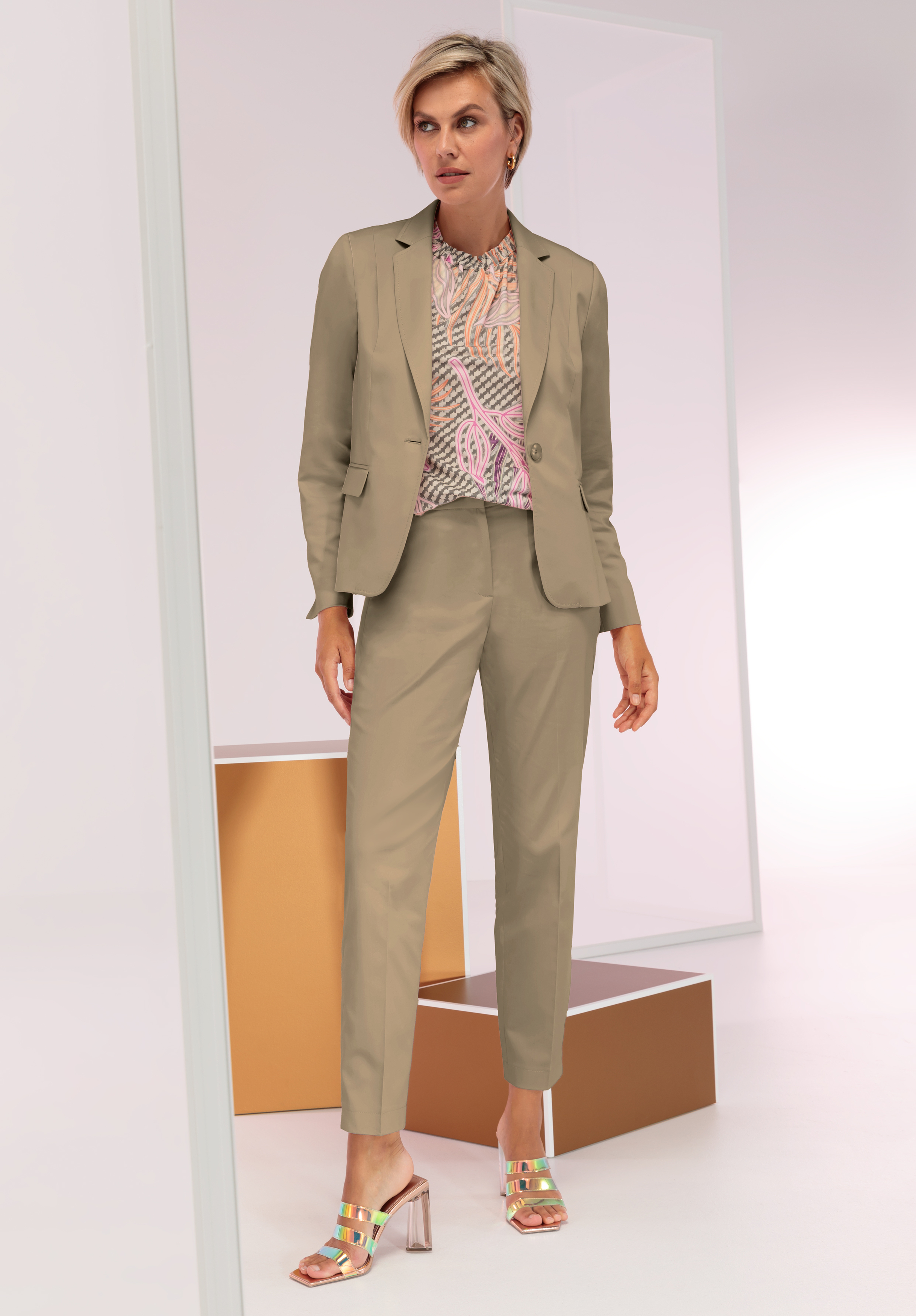 Eine Frau steht in einem hellbraunen Hosenanzug, einer gemusterten Bluse und metallischen Pumps mit offenen Zehen. Sie hat kurze blonde Haare und posiert in einer minimalistischen Umgebung mit geometrischen Formen in Gold und Weiß.