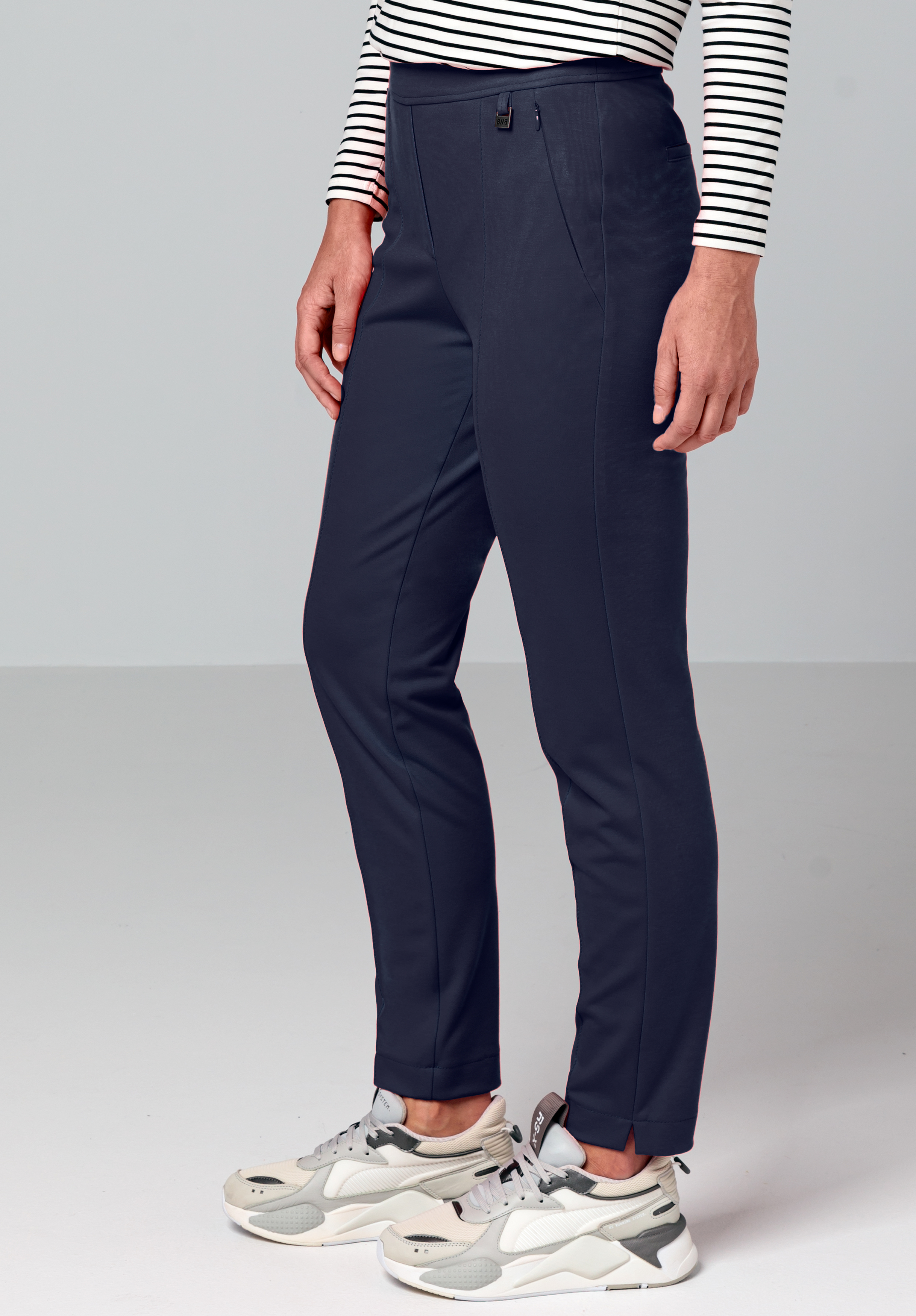 Eine Person, die ein weiß-schwarz gestreiftes Langarmoberteil, eine marineblaue Hose mit Vordernaht und weiße Turnschuhe trägt, steht vor einem schlichten hellen Hintergrund.