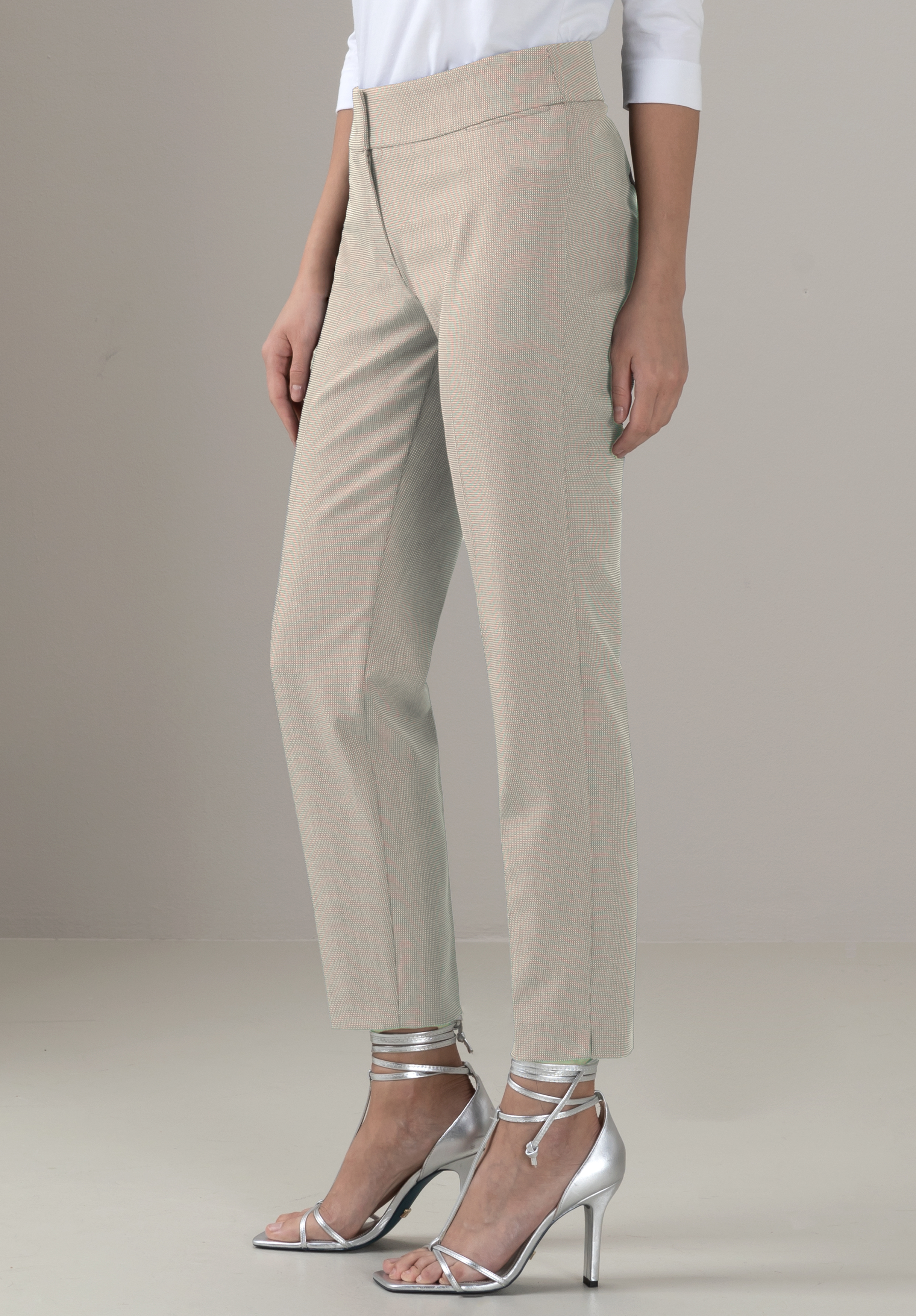 Eine Person, die eine hellbeige maßgeschneiderte Hose und ein weißes Oberteil trägt, steht in einem Haus. Sie trägt silberne hochhackige Sandalen mit Knöchelriemen. Der Hintergrund ist schlicht und neutral.