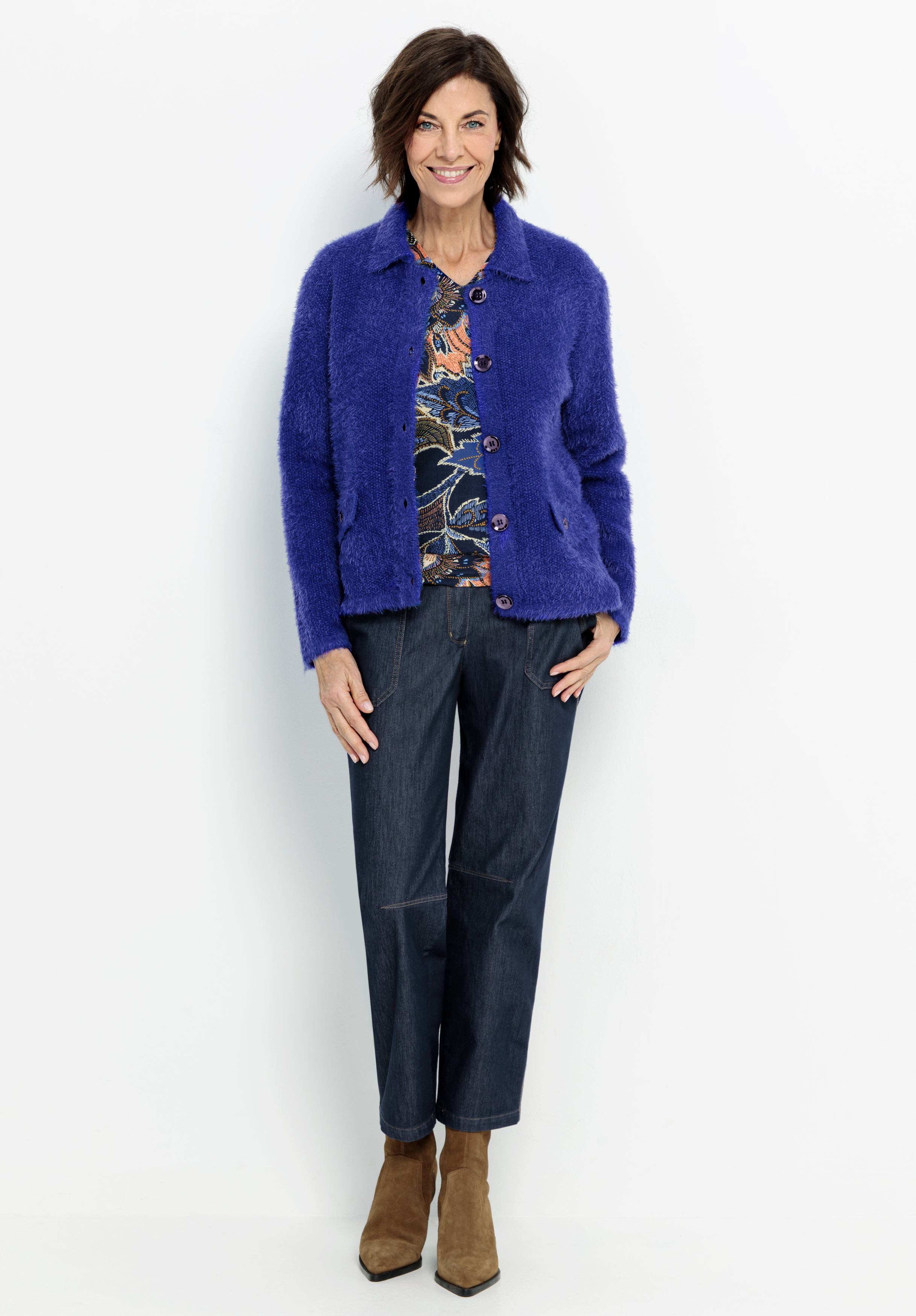 Eine Frau steht lächelnd vor einem weißen Hintergrund und trägt eine wuschelige lila Jacke über einer gemusterten Bluse, dunkelblaue Jeans und hellbraune Stiefeletten. Sie hat kurzes braunes Haar und ihre Hände sind an den Seiten.