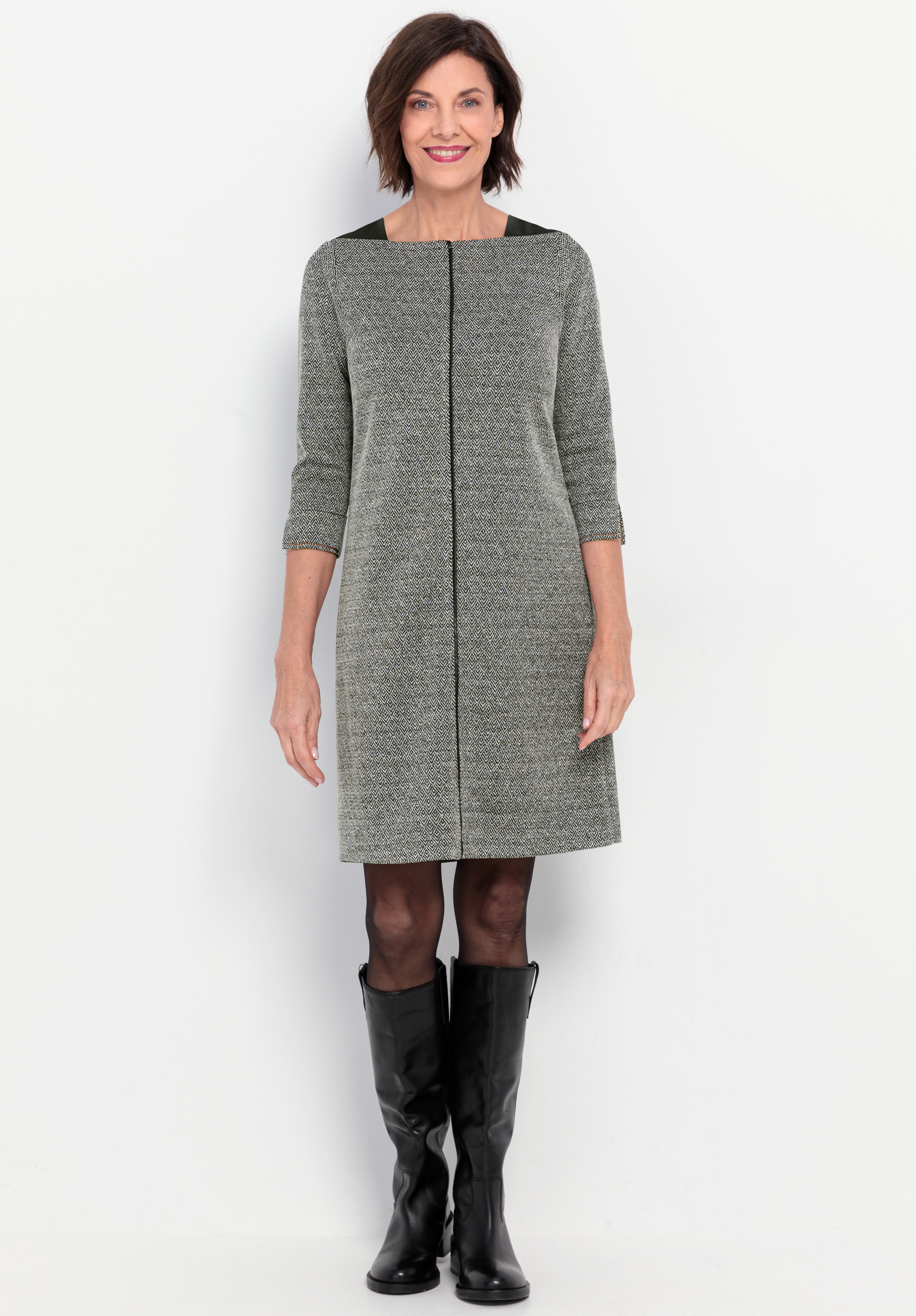 Eine Frau steht lächelnd vor einem weißen Hintergrund und trägt ein graues, knielanges Kleid mit Dreiviertelärmeln, eine schwarze Strumpfhose und schwarze, kniehohe Stiefel.