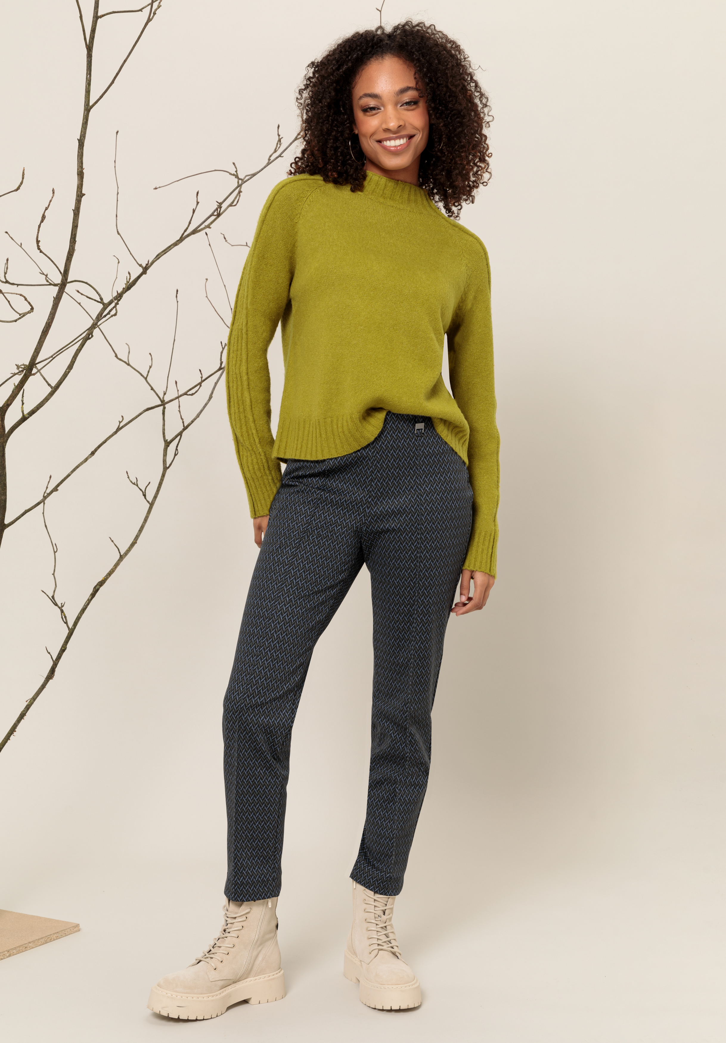Eine Frau mit lockigem Haar, die einen grünen Pullover, eine dunkel gemusterte Hose und hellbeige Schnürstiefel trägt, steht lächelnd vor einem neutralen Hintergrund mit einer Dekoration aus kahlen Zweigen neben sich.