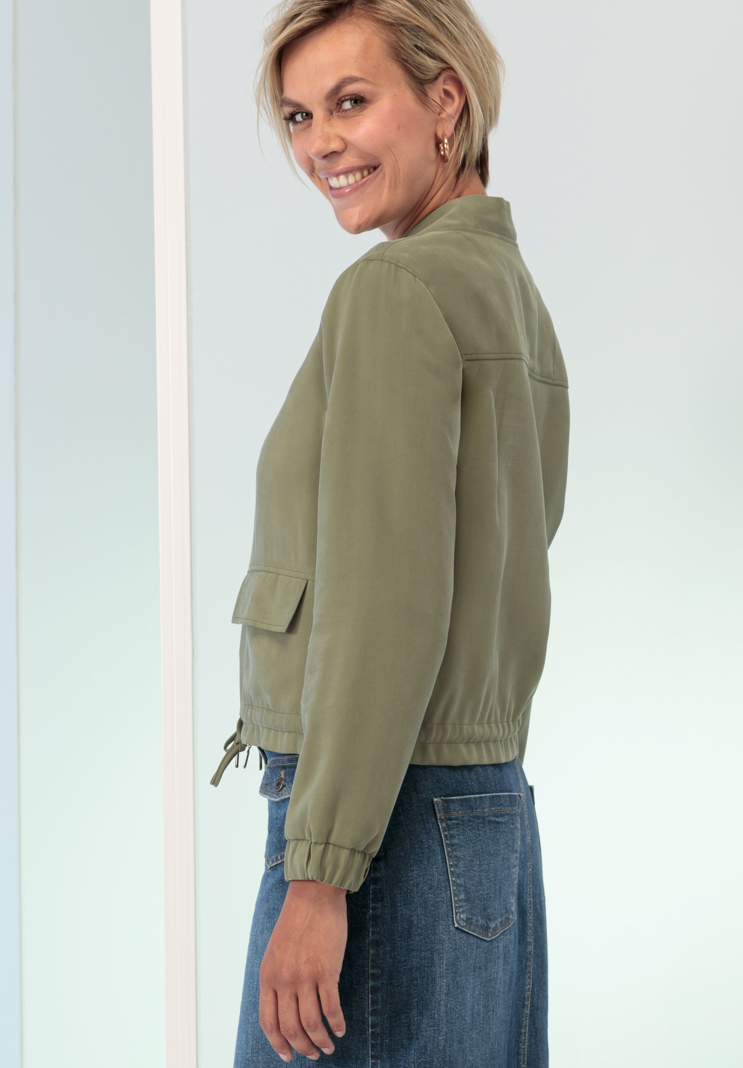 Eine Frau mit kurzen blonden Haaren, die eine hell olivgrüne Jacke und einen blauen Jeansrock trägt, steht seitlich und lächelt vor einem schlichten, hellen Hintergrund in die Kamera.