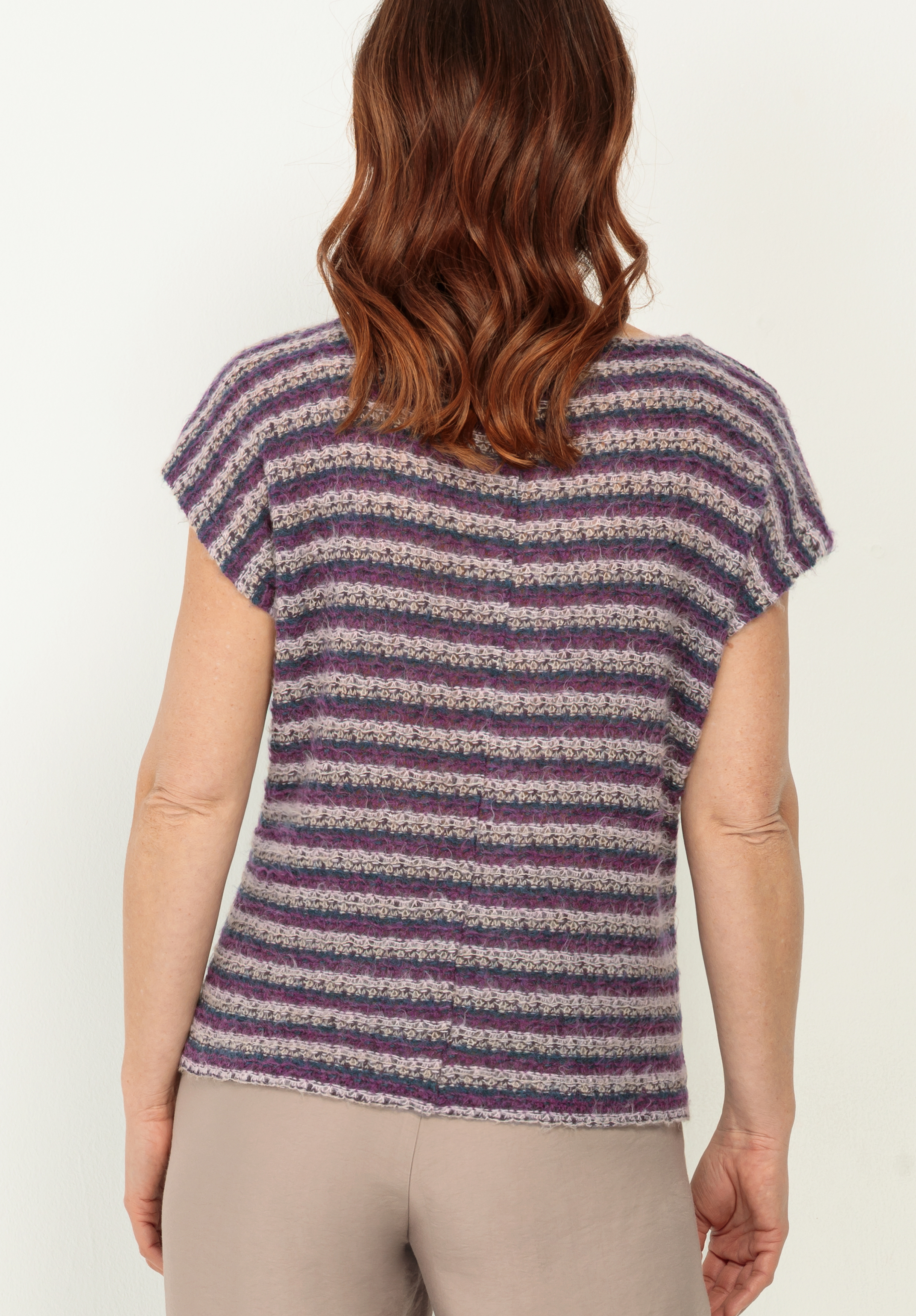Eine Frau mit gewelltem, schulterlangem braunem Haar ist von hinten zu sehen. Sie trägt ein kurzärmeliges, lila und grau gestreiftes Strickoberteil und eine hellbeige Hose und steht vor einem schlichten weißen Hintergrund.