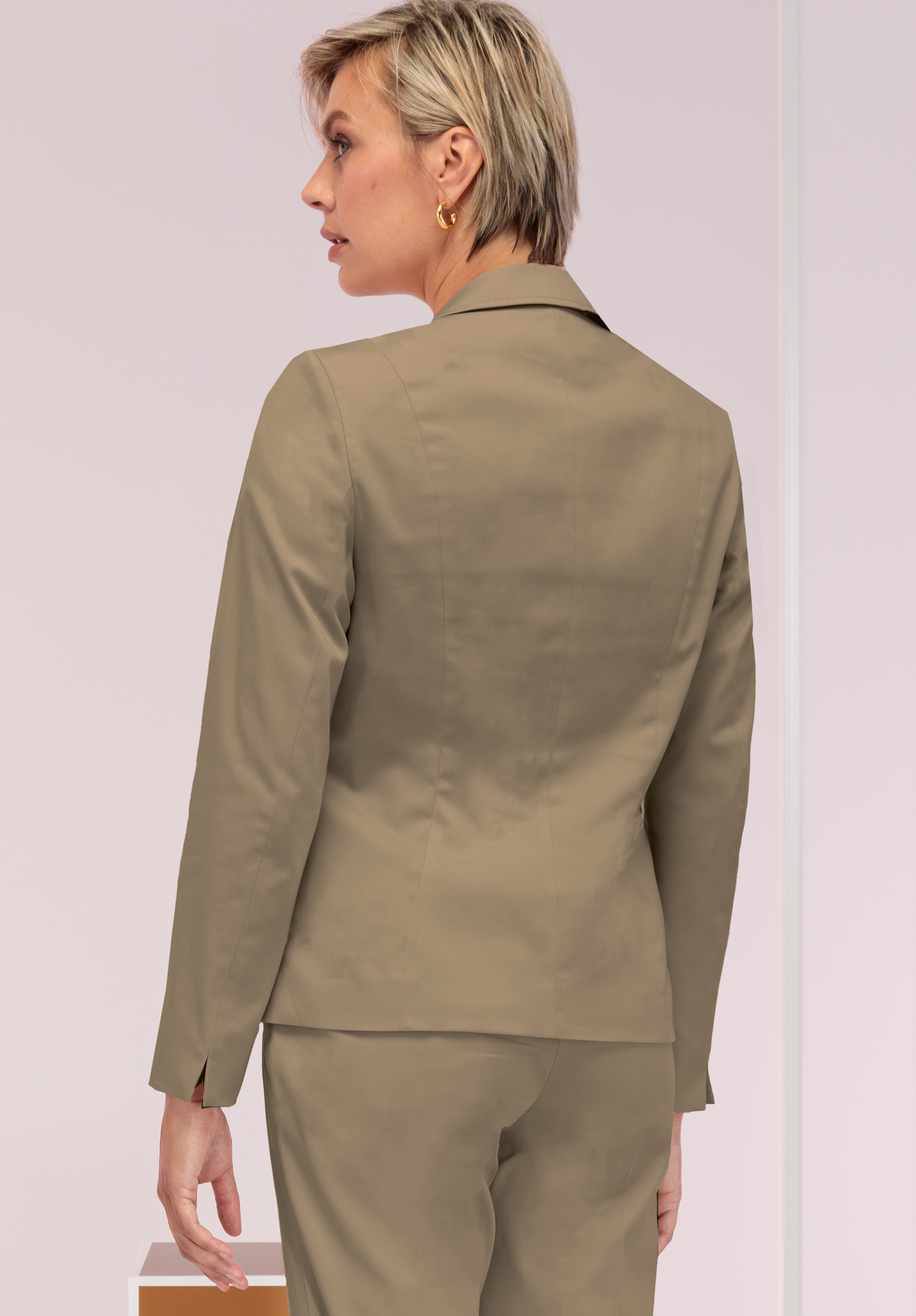 Eine Person mit kurzen blonden Haaren, die einen hellbraunen Blazer und eine passende Hose trägt, steht mit dem Rücken zur Kamera in einer minimalistischen Umgebung mit hellem Hintergrund.