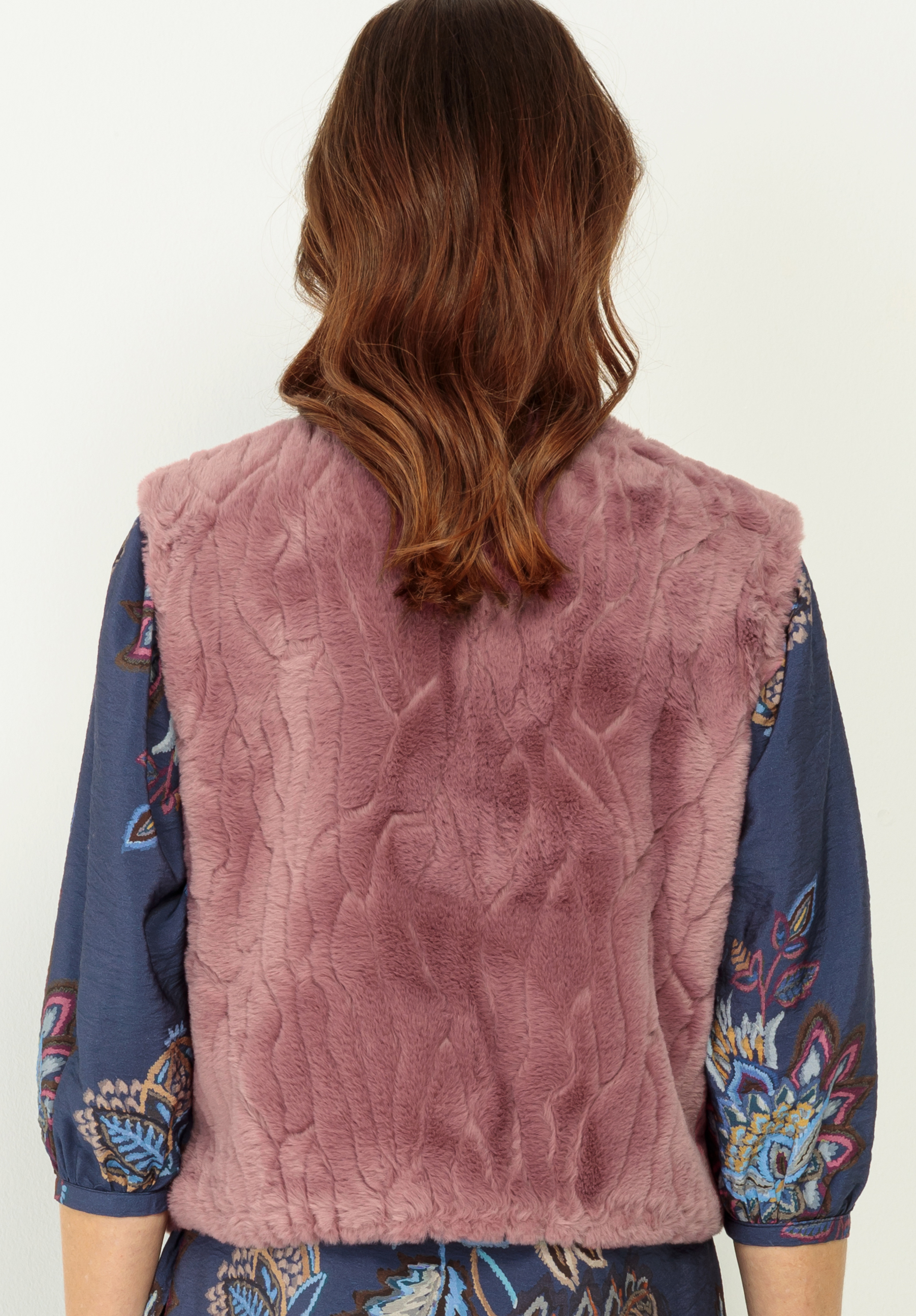 Eine Person mit gewelltem braunem Haar, von hinten gesehen, trägt eine strukturierte, lila-rosafarbene Kunstpelzweste über einem marineblauen Oberteil mit einem bunten Blumenmuster.