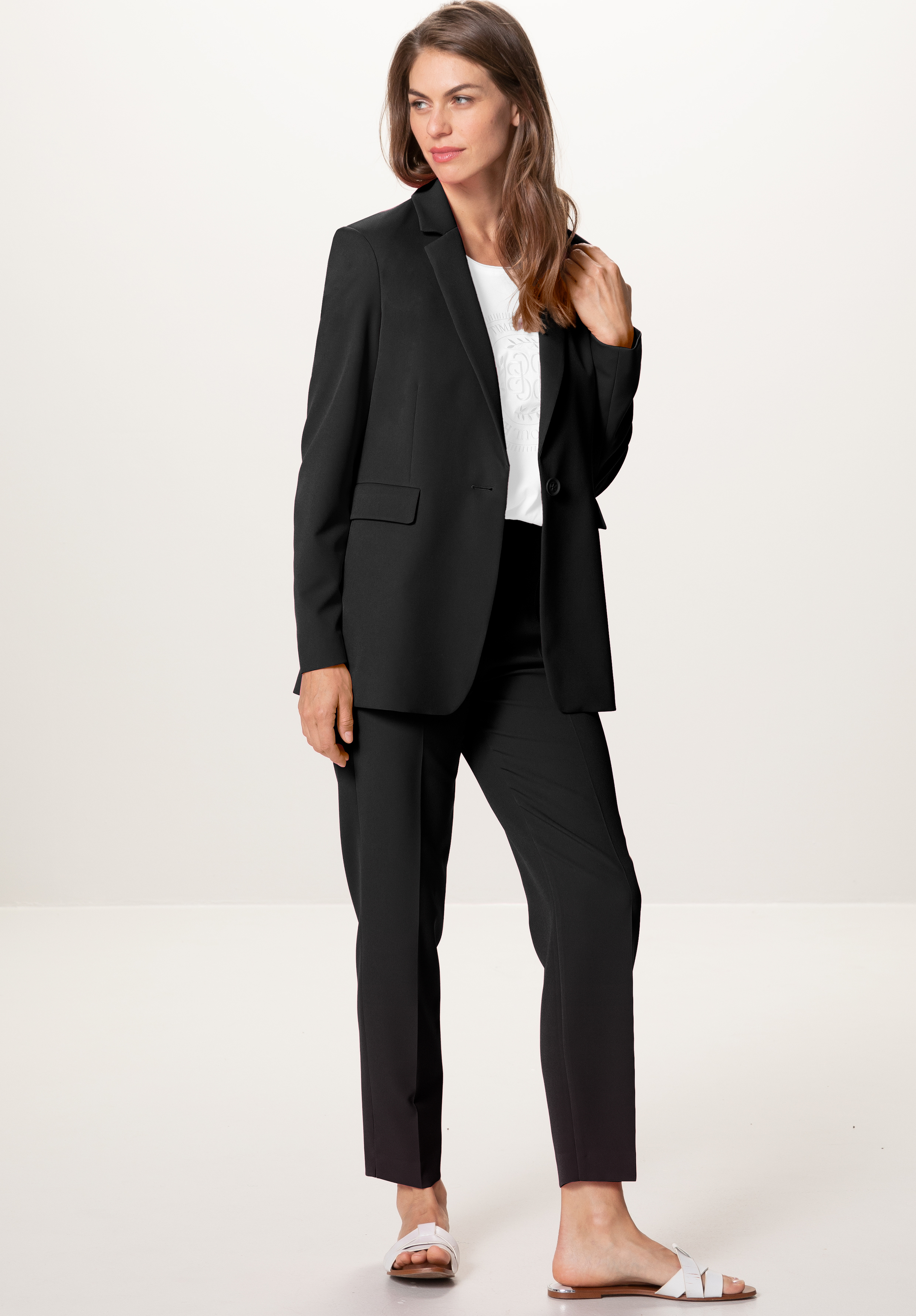 Eine Frau steht vor einem schlichten Hintergrund und trägt einen schwarzen Anzug mit einem weißen Oberteil und weißen Slide-Sandalen. Sie hat langes braunes Haar, ein leichtes Lächeln, und ihre linke Hand berührt ihren Blazerkragen.