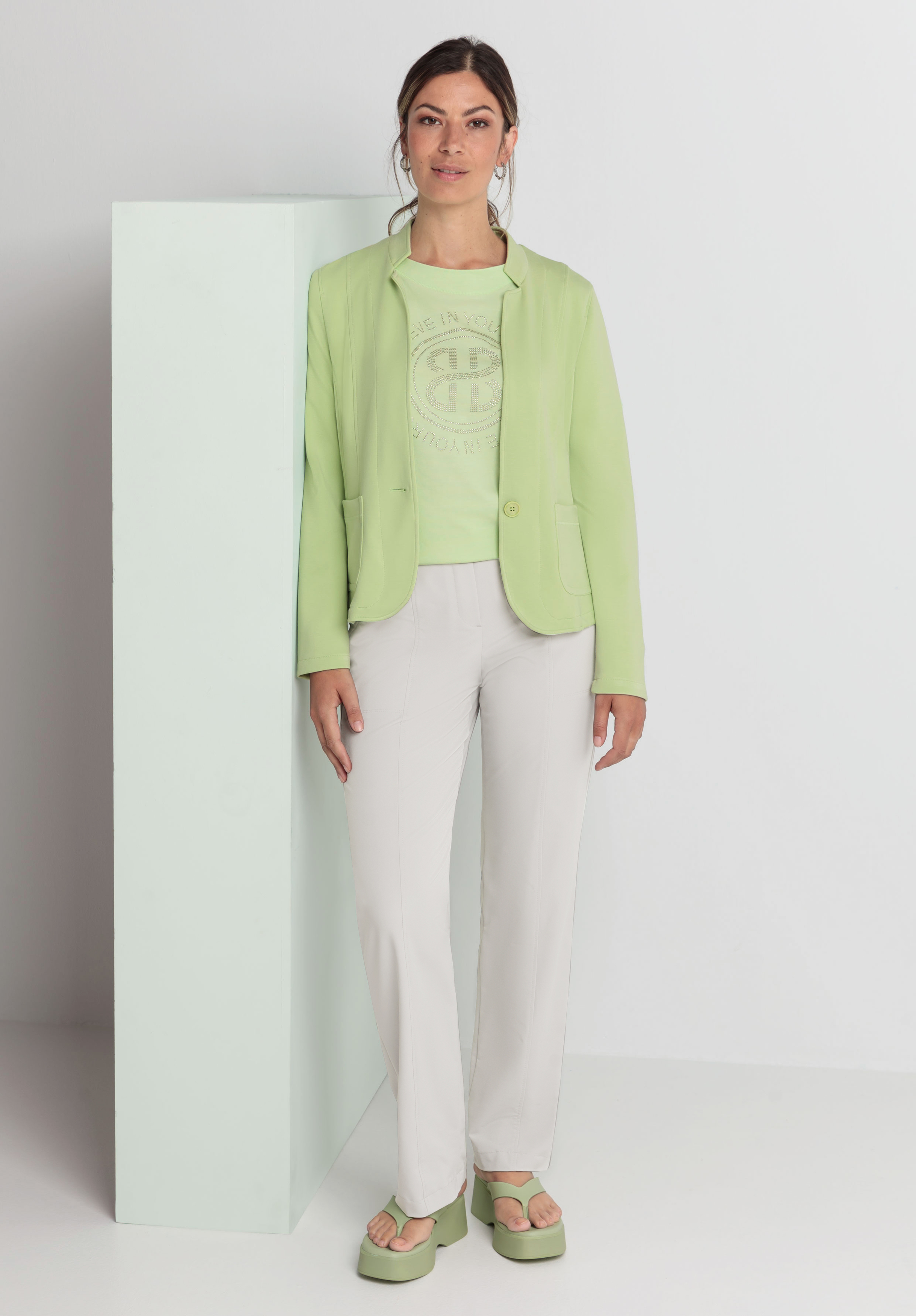 Eine Frau steht vor einem hellen Hintergrund. Sie trägt einen hellgrünen Blazer über einem passenden T-Shirt, eine weiße Hose und hellgrüne Plateausandalen. Sie hat ihre Haare zurückgebunden und steht neben einem hellmintfarbenen rechteckigen Block.