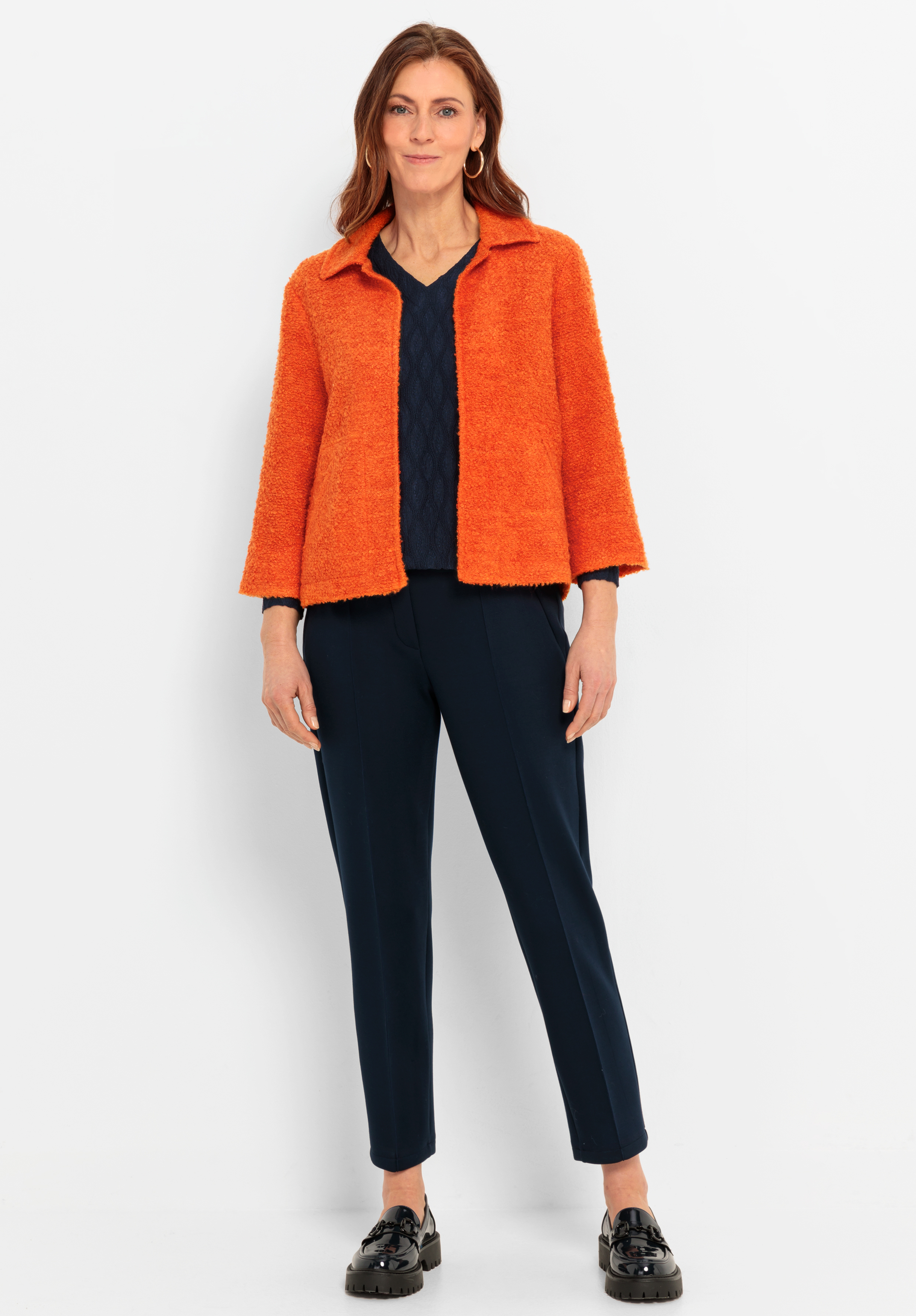 Eine Frau trägt eine strukturierte orangefarbene Jacke über einem dunkelblauen Oberteil und eine marineblaue Hose, gepaart mit schwarzen Halbschuhen. Sie hat langes braunes Haar.