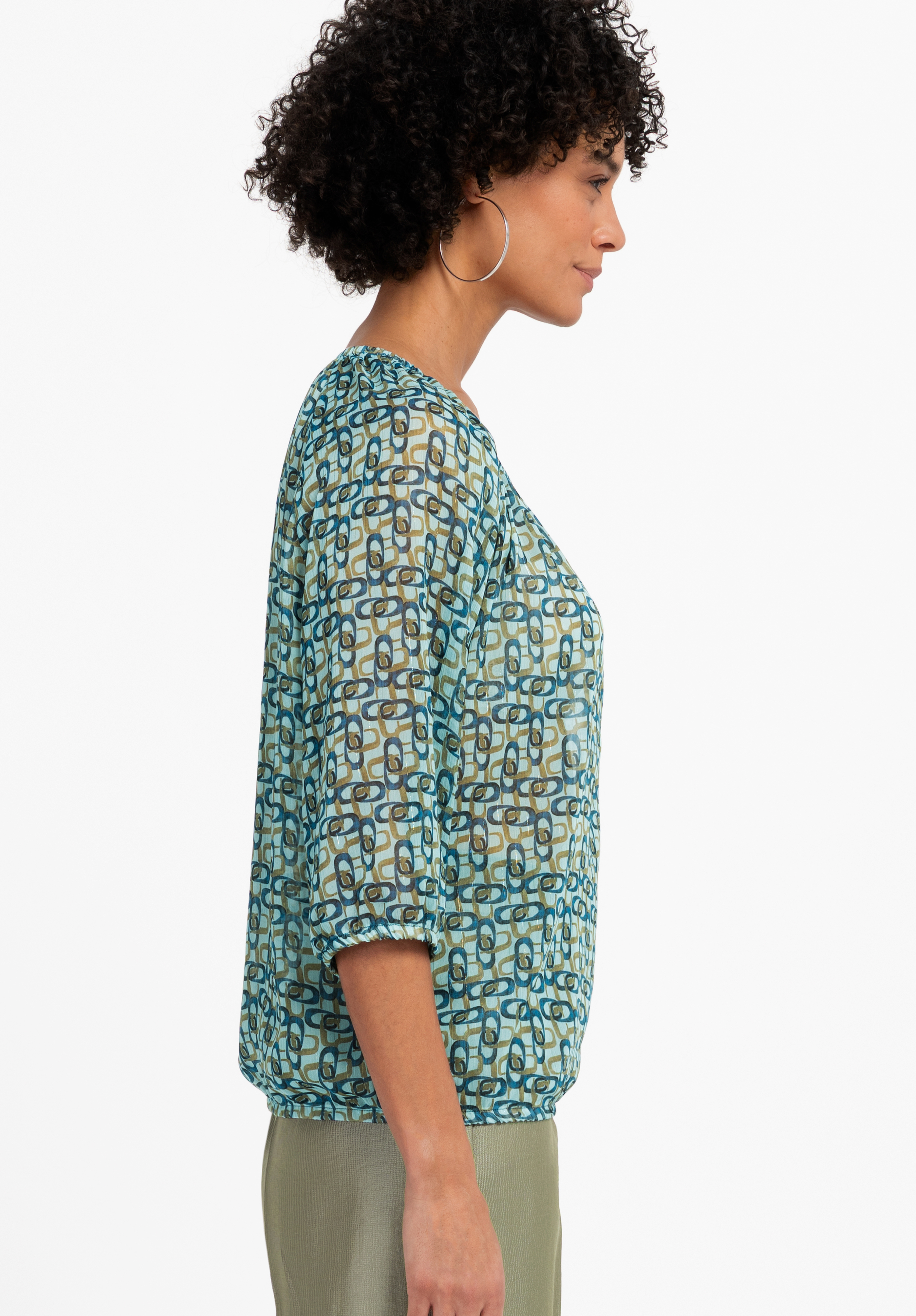 Eine Frau mit lockigem Haar trägt ein hellblaues Langarmshirt mit weißen und grünen Längsstreifen und blaue Jeans und blickt vor einem schlichten Hintergrund über ihre Schulter.