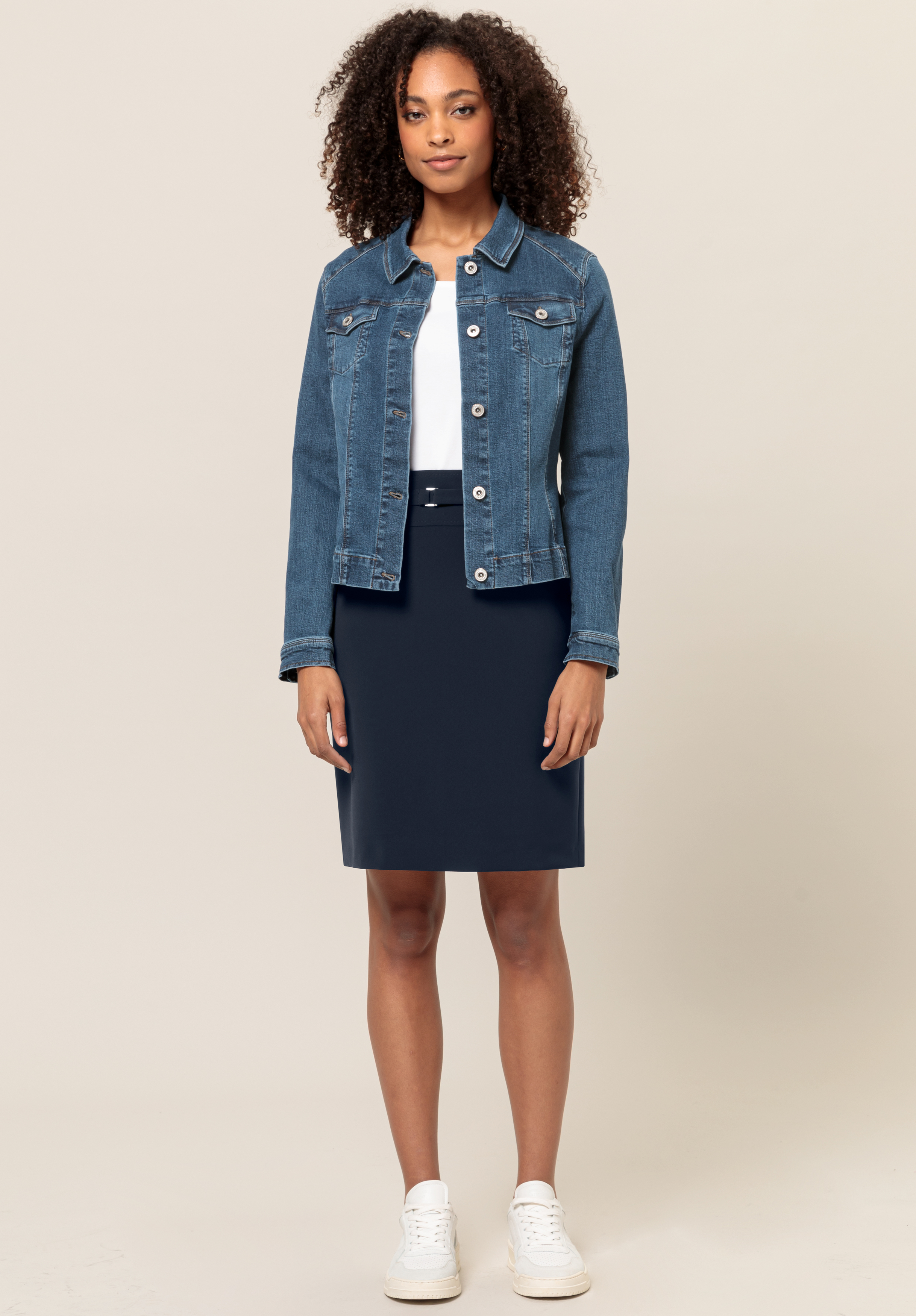 Eine Frau steht mit dem Gesicht nach vorne, trägt eine blaue Jeansjacke über einem weißen Oberteil, einen marineblauen knielangen Rock und weiße Turnschuhe vor einem schlichten beigen Hintergrund.