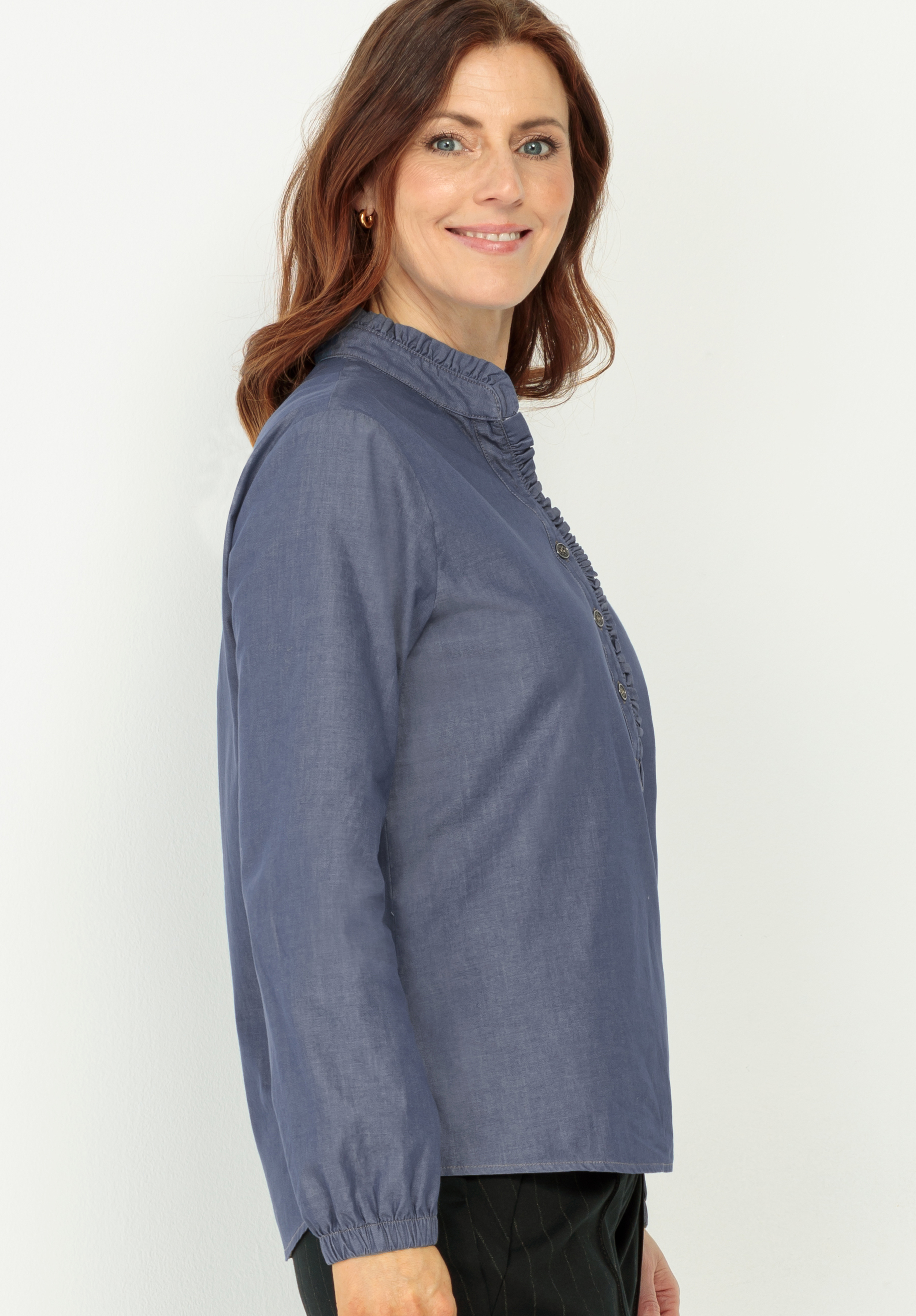 Eine Frau mit schulterlangen braunen Haaren und blauen Augen steht lächelnd seitlich und blickt nach links. Sie trägt eine langärmelige, blaue Bluse mit Rüschenkragen und eine schwarze Hose. Der Hintergrund ist einfarbig weiß.
