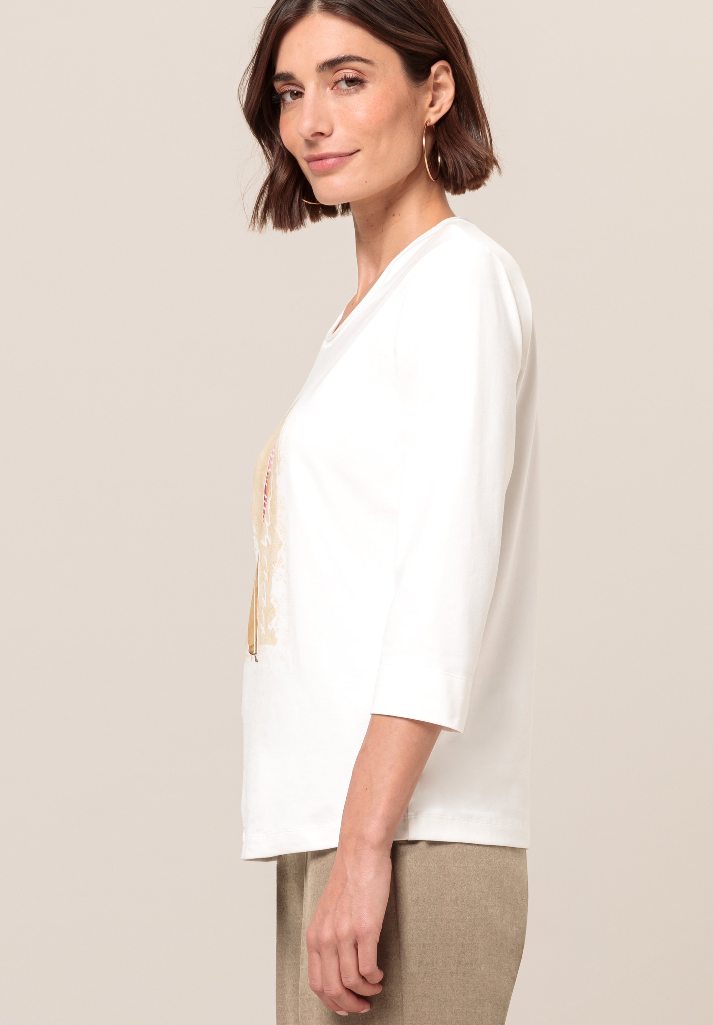 Frau mit kurzen braunen Haaren, in einem weißen Oberteil mit Dreiviertelärmeln und beigen Hosen, steht seitlich und lächelt sanft vor einem schlichten hellen Hintergrund.