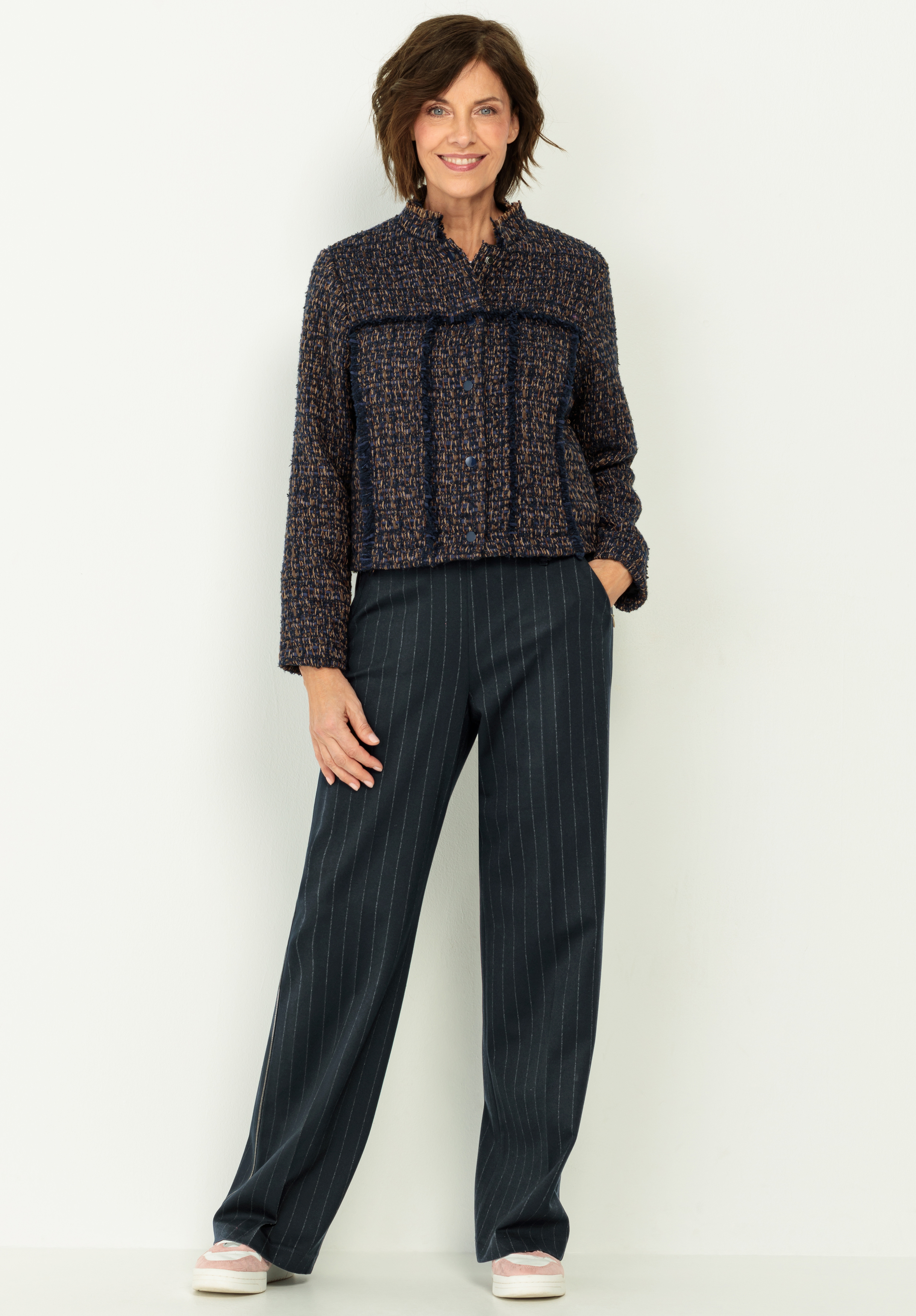 Eine Frau steht lächelnd vor einem hellen Hintergrund und trägt eine strukturierte dunkle Jacke mit Krawatte und eine lockere dunkle Nadelstreifenhose. Sie hat eine Hand in ihrer Tasche und trägt weiße Turnschuhe.