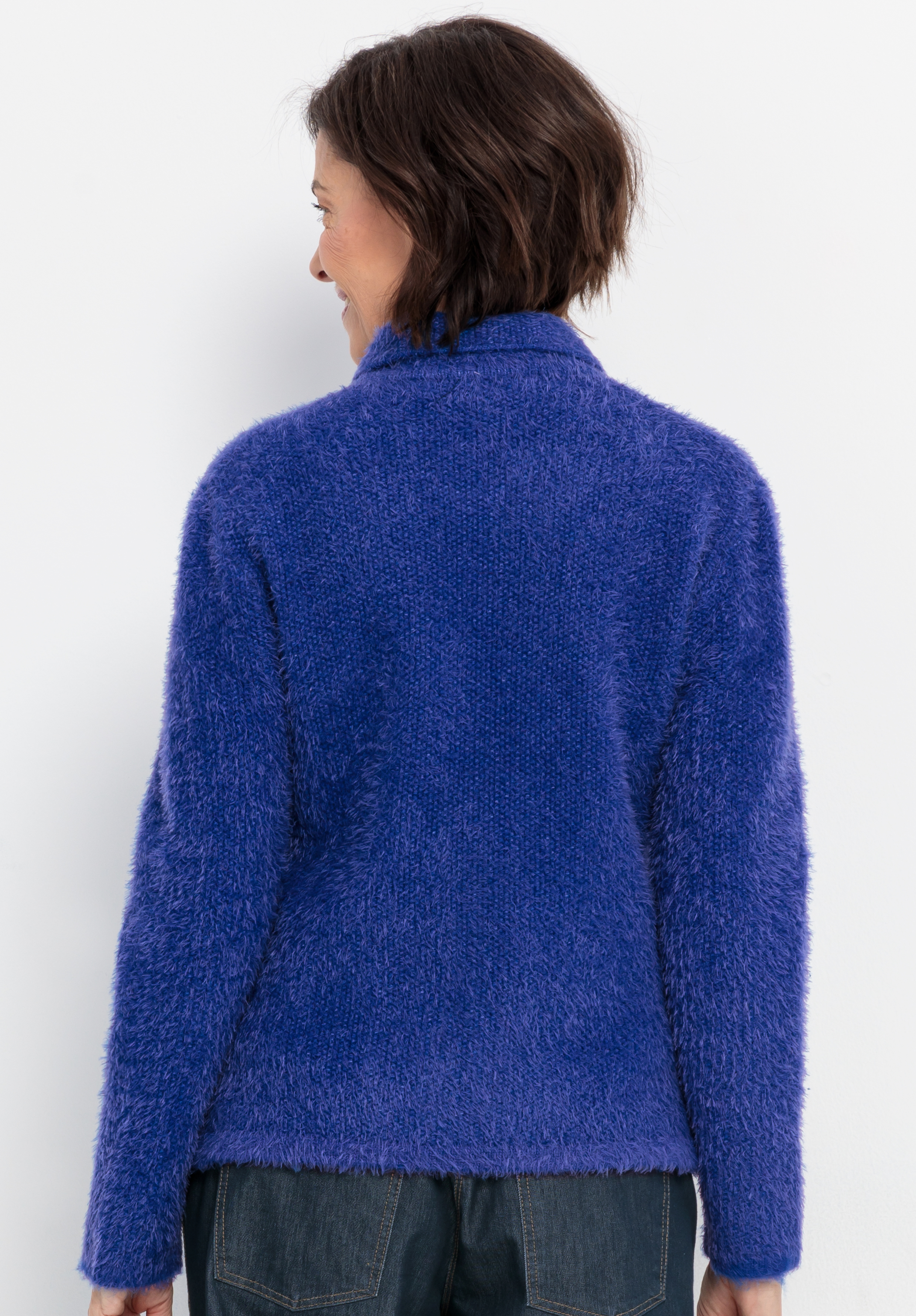 Eine Person mit kurzen braunen Haaren steht mit dem Rücken zur Kamera, trägt einen weichen, flauschigen königsblauen Pullover und dunkle Jeans vor einem schlichten weißen Hintergrund.