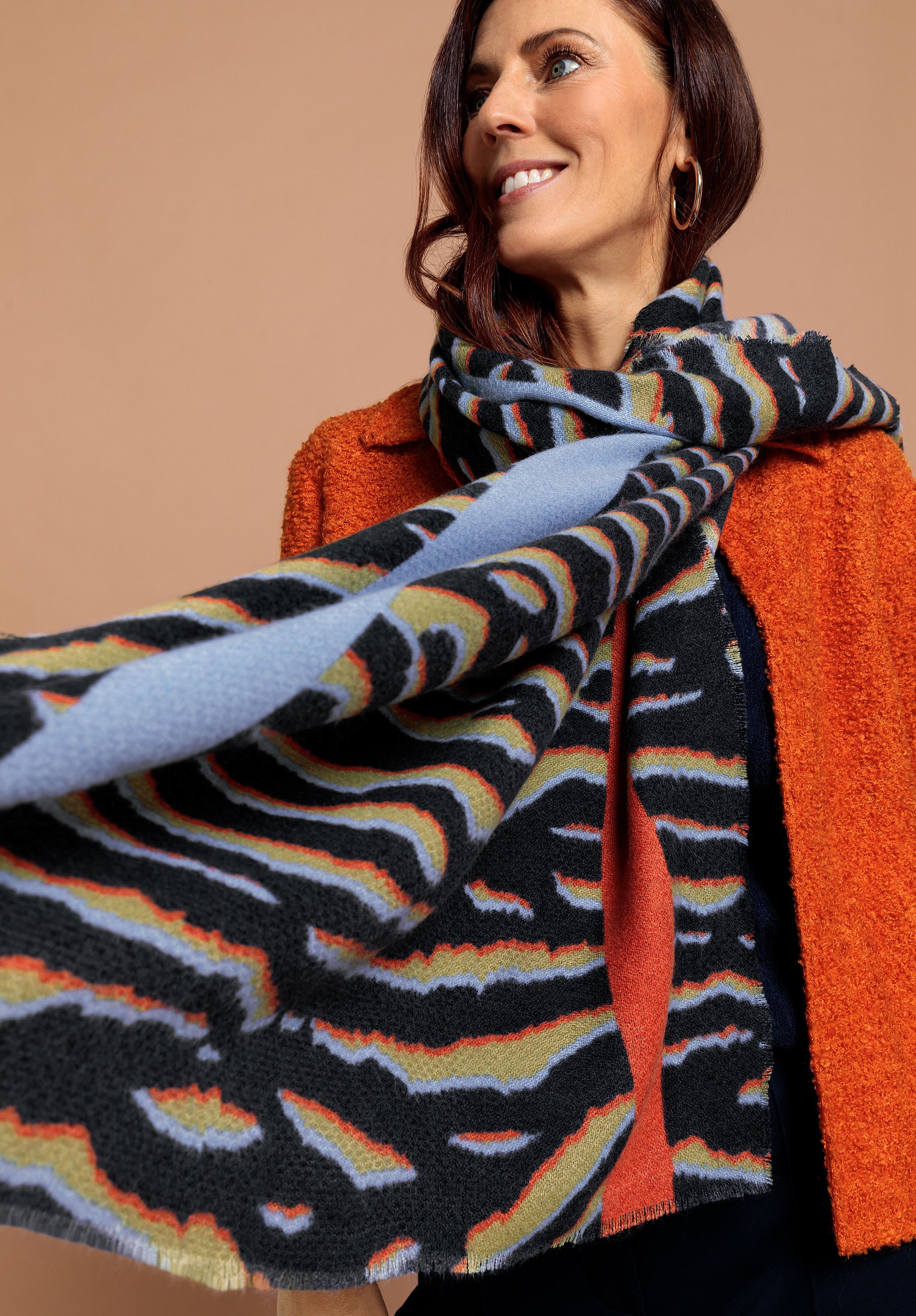 Modeschal TORO Eine Frau, die eine orangefarbene, strukturierte Jacke und einen großen, bunt gemusterten Schal trägt, lächelt, während sie zur Seite schaut, und steht vor einem schlichten, hellbraunen Hintergrund.