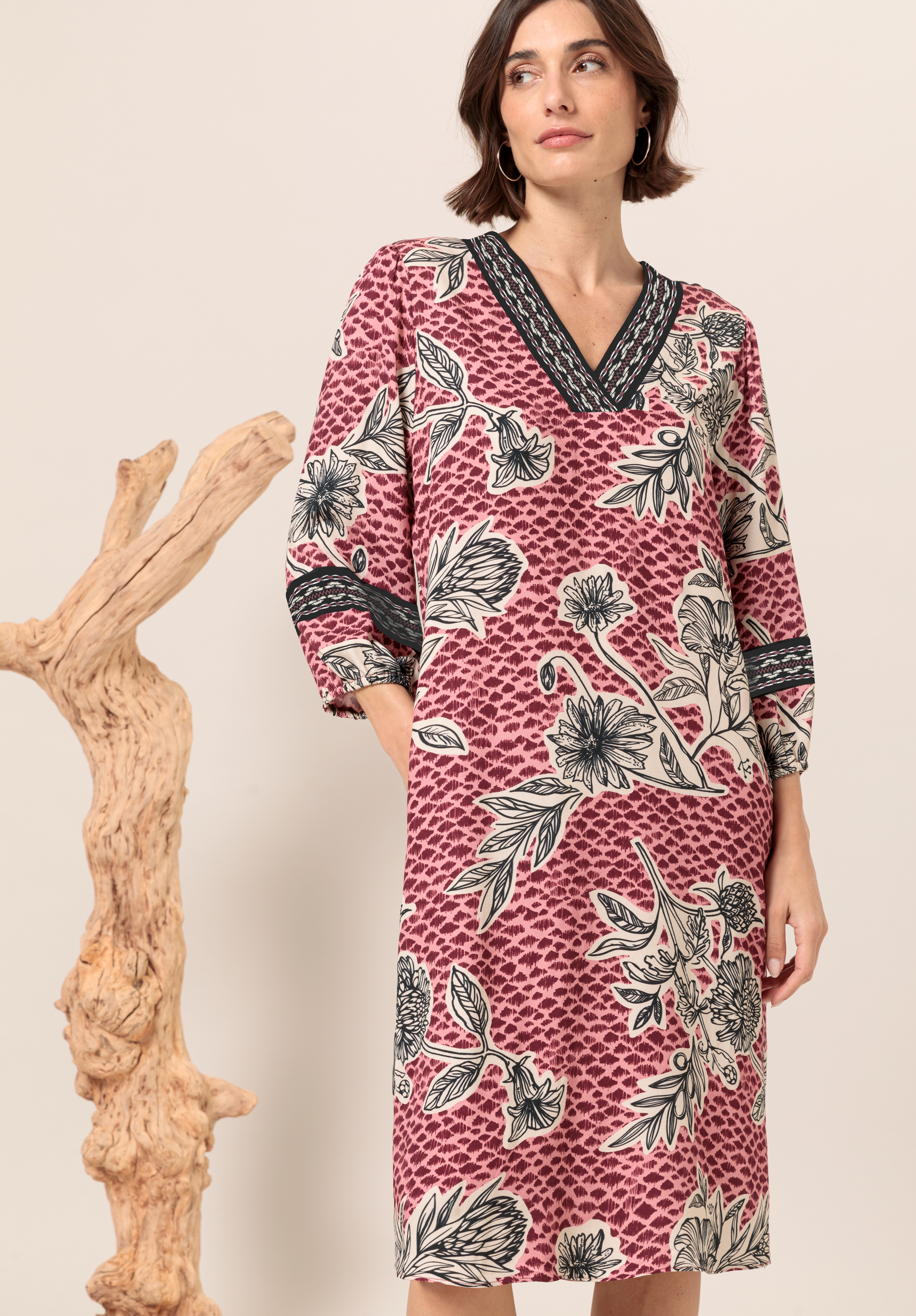 Druckkleid ALESSA Eine Frau in einem knielangen, langärmeligen, rosa Kleid mit schwarz-weißem Blumenmuster steht neben einem skulpturalen Stück aus hellem Holz, blickt leicht zur Seite und hat eine Hand in der Tasche.