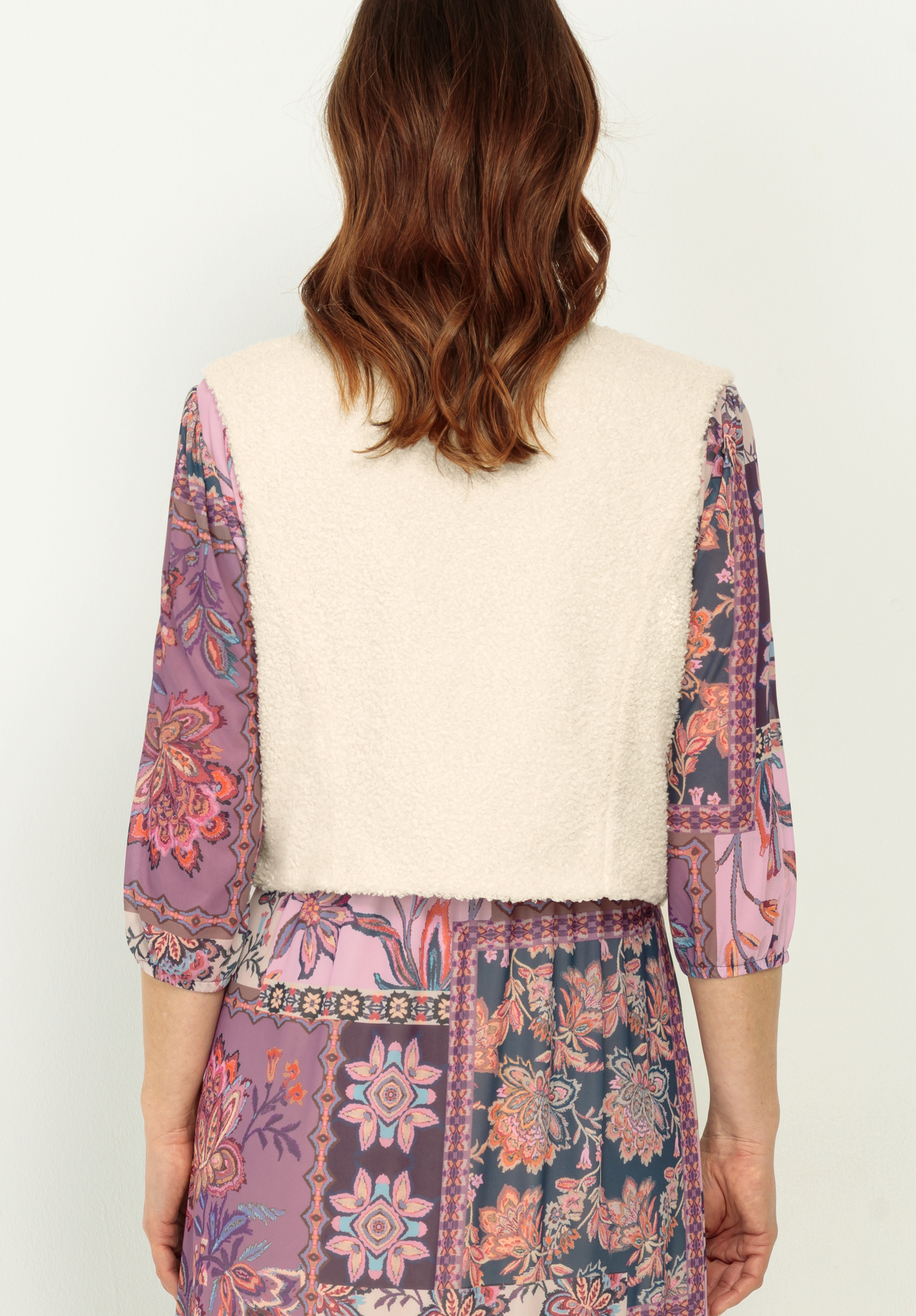 Eine Person mit gewelltem braunem Haar trägt eine cremefarbene, strukturierte Weste über einem langärmeligen Kleid mit einem lila, rosa und blaugrünen Blumenflickenmuster (von hinten gesehen).