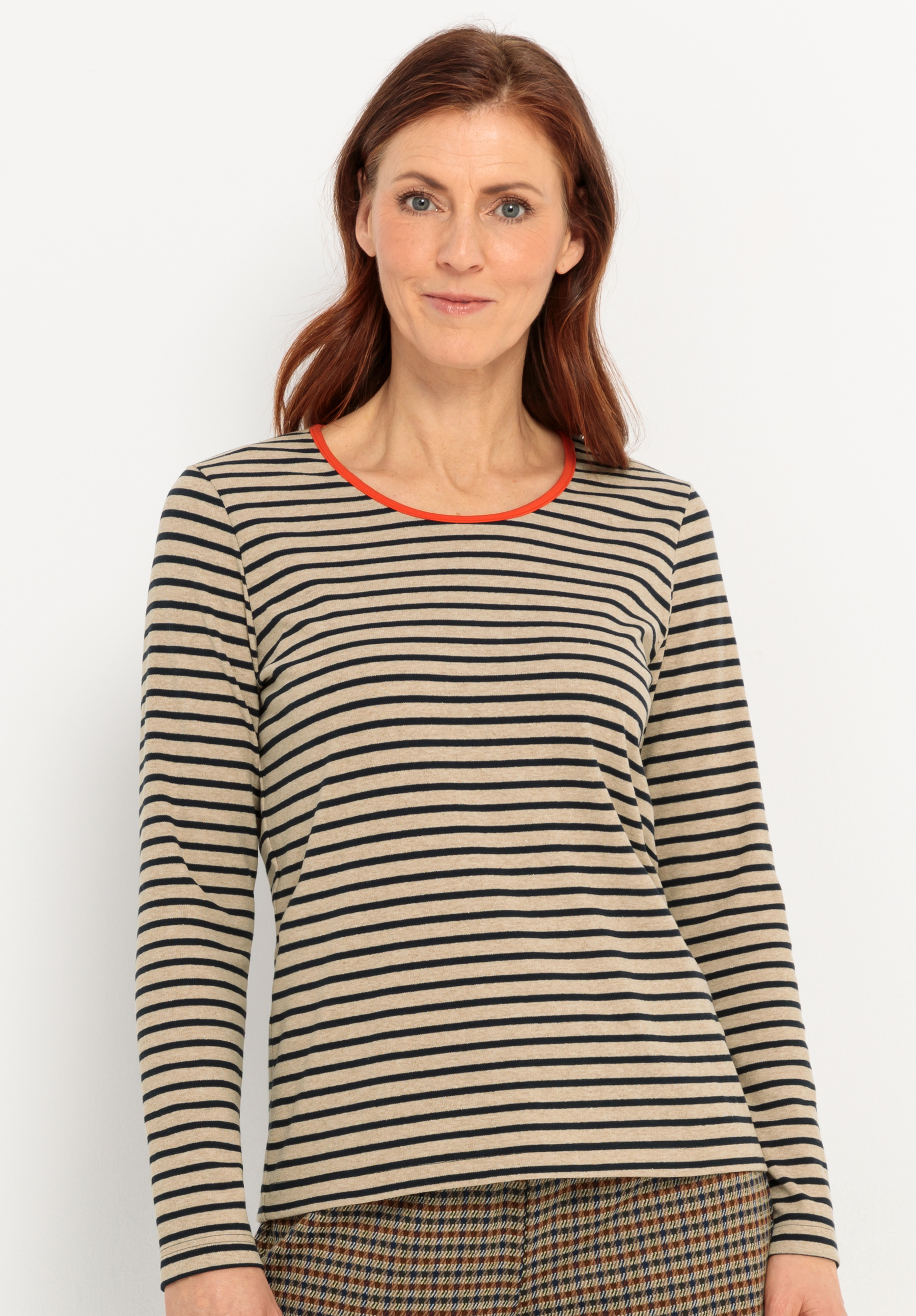 Langarmshirt DAMI Eine Frau mit langen braunen Haaren, die ein beige-schwarz gestreiftes langärmeliges Hemd mit rotem Ausschnitt und eine karierte Hose trägt, steht vor einem schlichten weißen Hintergrund und lächelt sanft.
