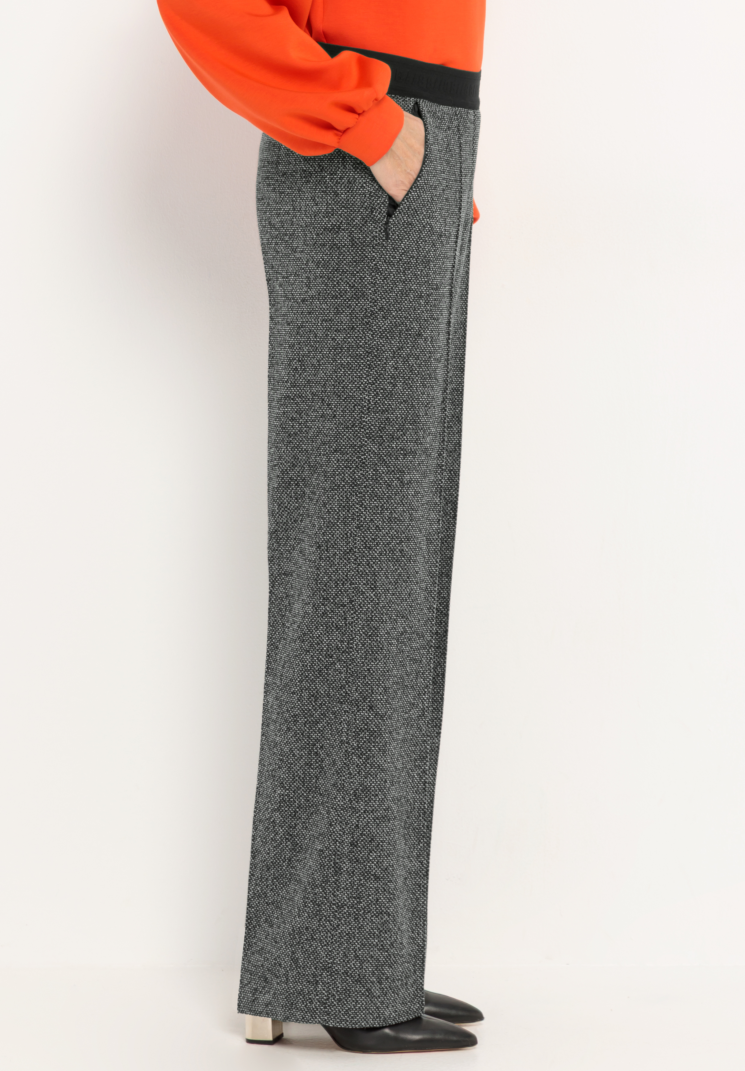 Eine Person, die eine graue Hose mit weitem Bein, ein leuchtend orangefarbenes Langarm-Oberteil und schwarze Schuhe mit spitzer Zehenpartie trägt und seitlich vor einem schlichten weißen Hintergrund steht.