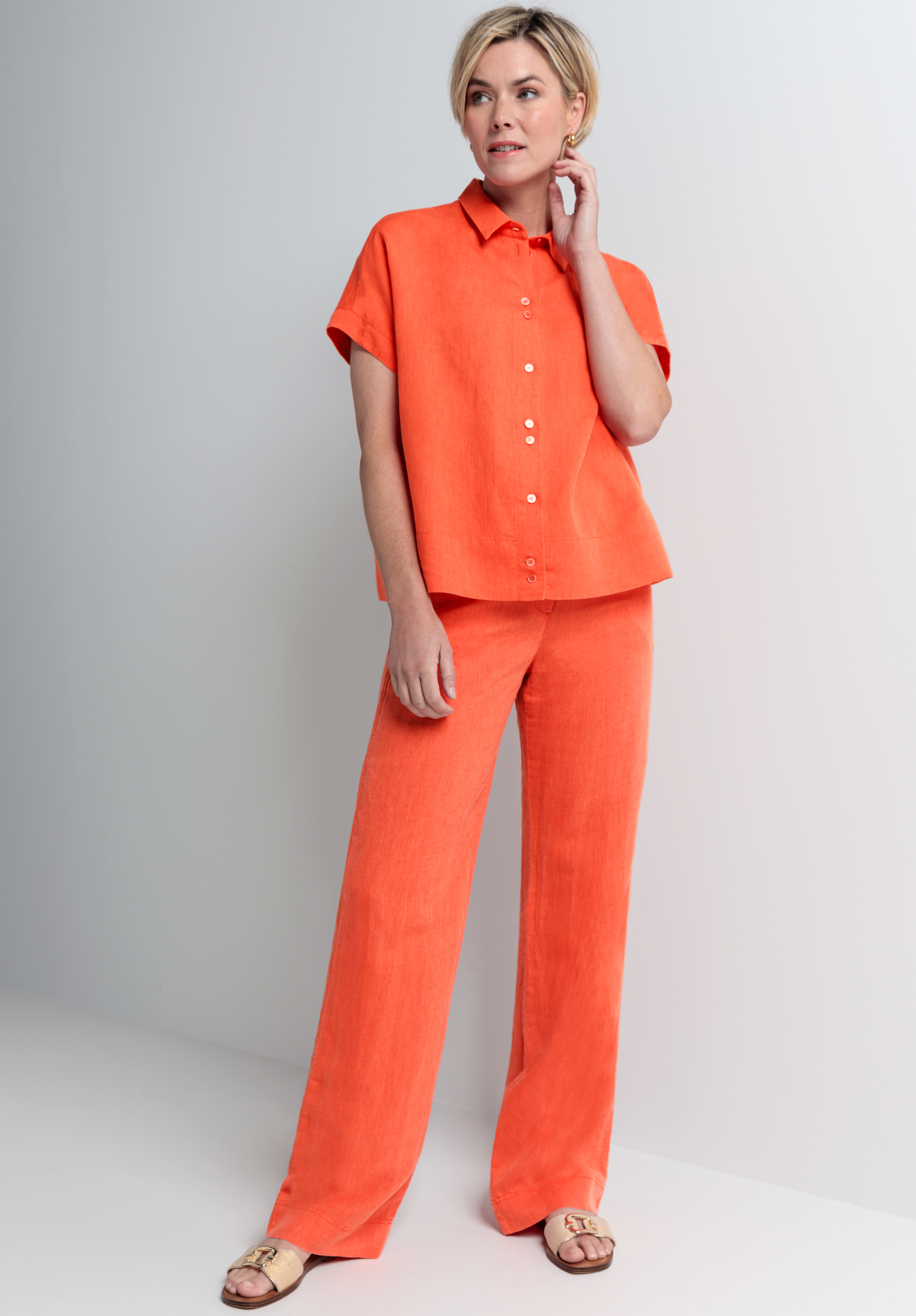 Eine Frau steht in einem minimalistisch gestalteten Raum. Sie trägt ein locker sitzendes, leuchtend orangefarbenes Kurzarmhemd, dazu passende weite Hosen und hellbraune Sandalen. Sie hat kurze blonde Haare, berührt ihren Hals und blickt leicht zur Seite.