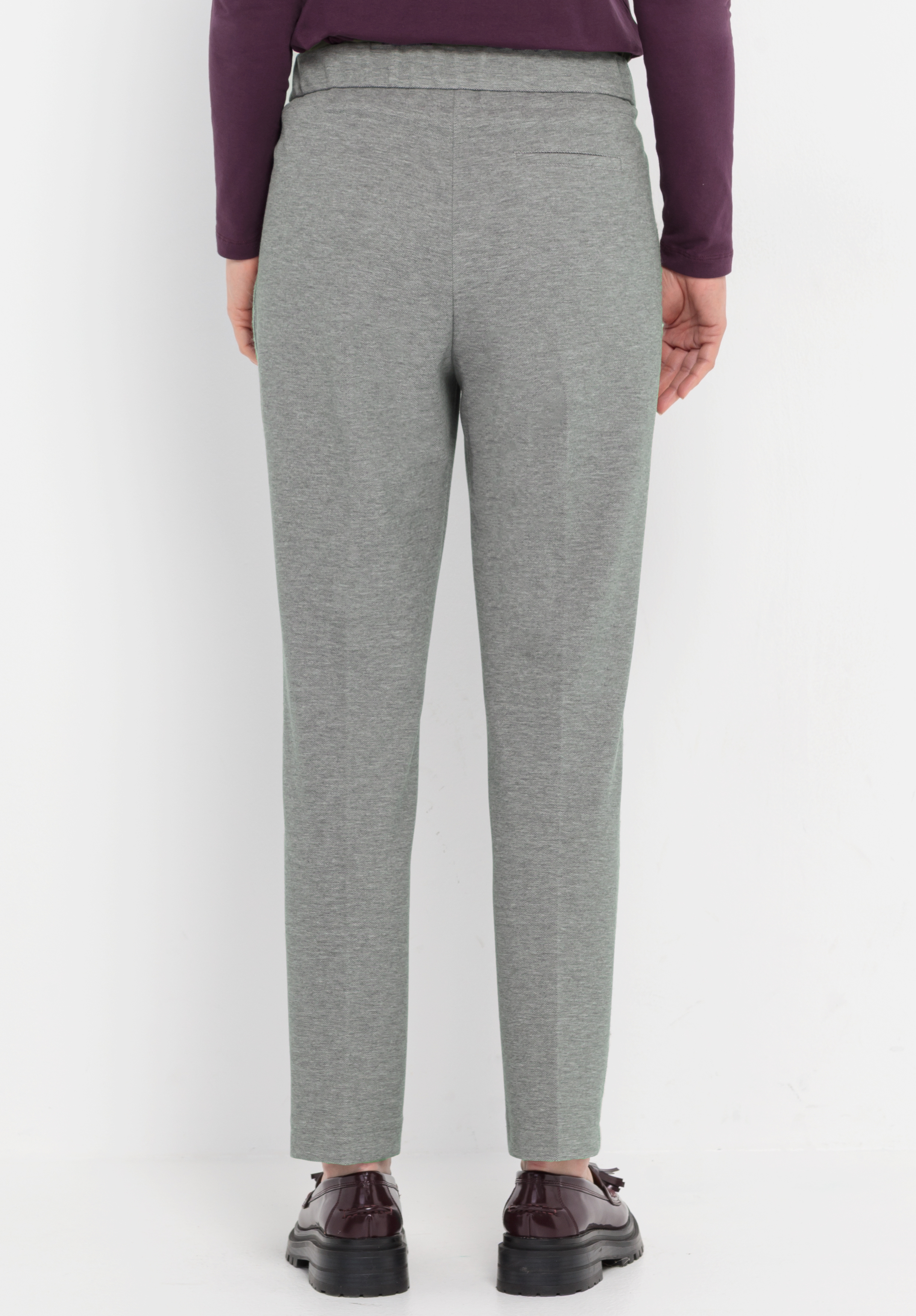 Eine Person, die eine graue, eng anliegende Hose, ein langärmeliges lilafarbenes Oberteil und dunkelbraune Schuhe trägt, steht vor einem schlichten weißen Hintergrund, von hinten gesehen.