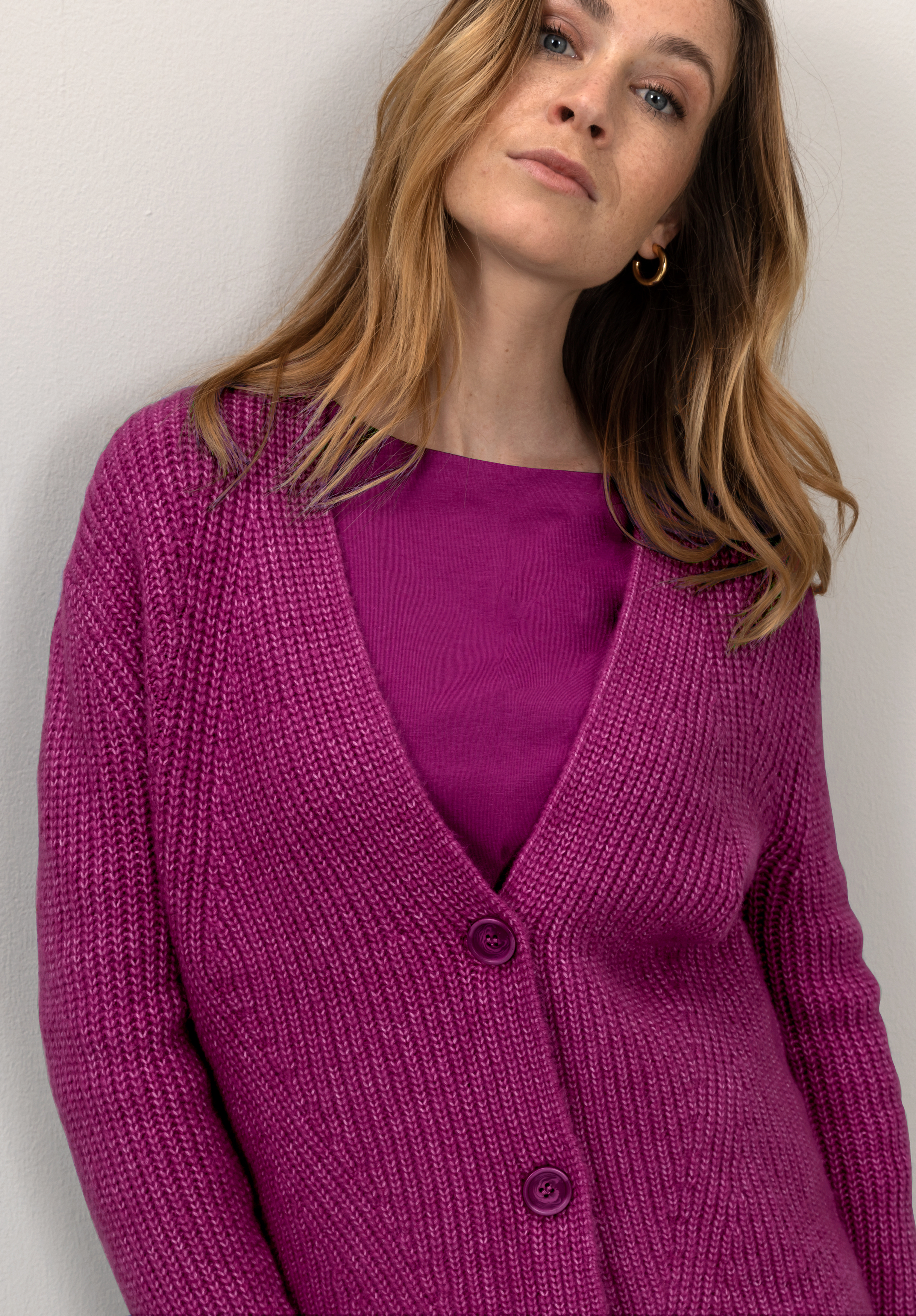 Eine Frau mit hellbraunem Haar trägt eine magentafarbene Strickjacke über einem dazu passenden magentafarbenen Oberteil, steht vor einer hellen Wand und blickt leicht nach oben in die Kamera.