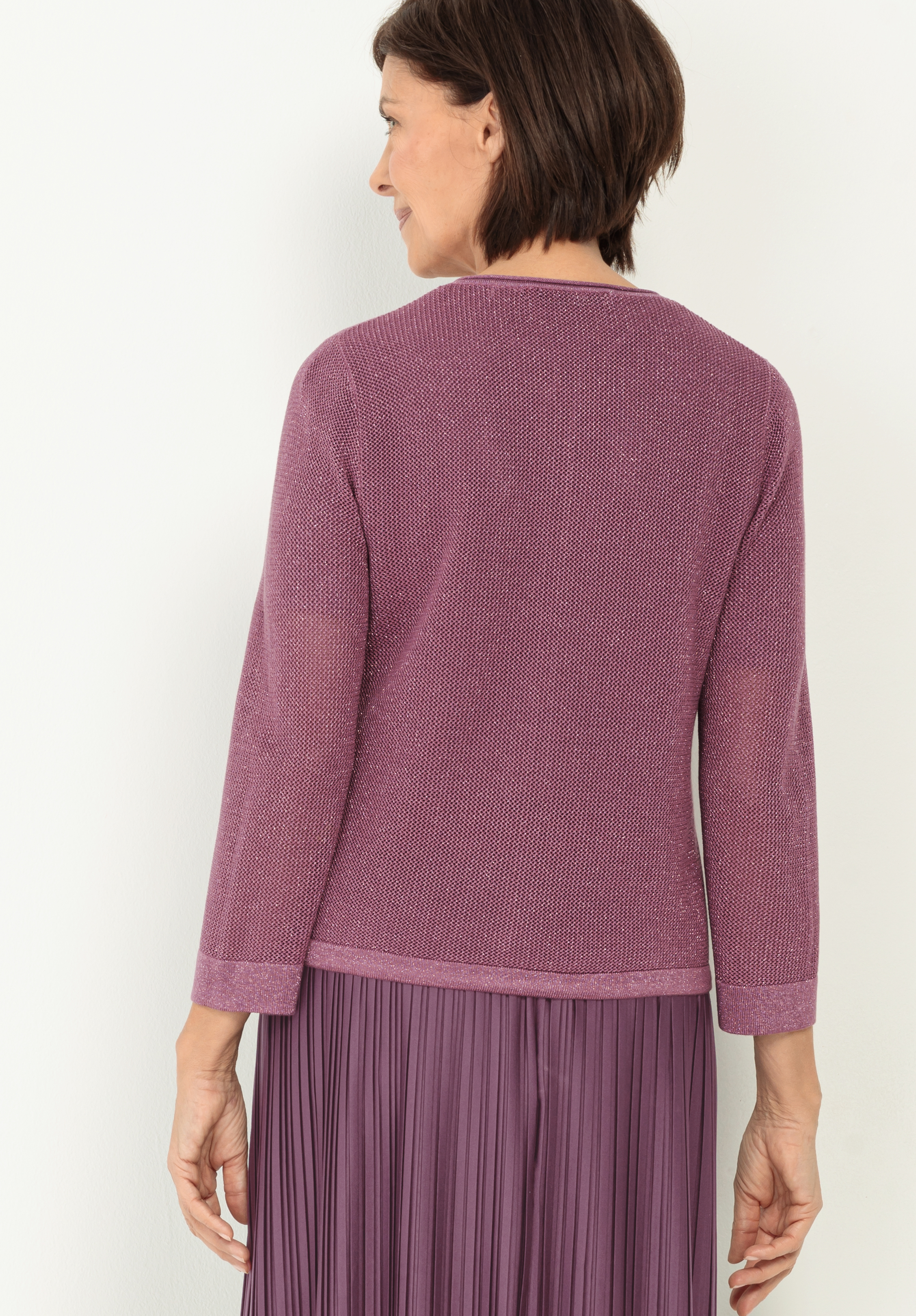 Eine Frau mit kurzen braunen Haaren, die einen lilafarbenen Strickpullover mit Struktur und einen lila Faltenrock trägt, steht vor einem hellen Hintergrund von der Kamera weg.