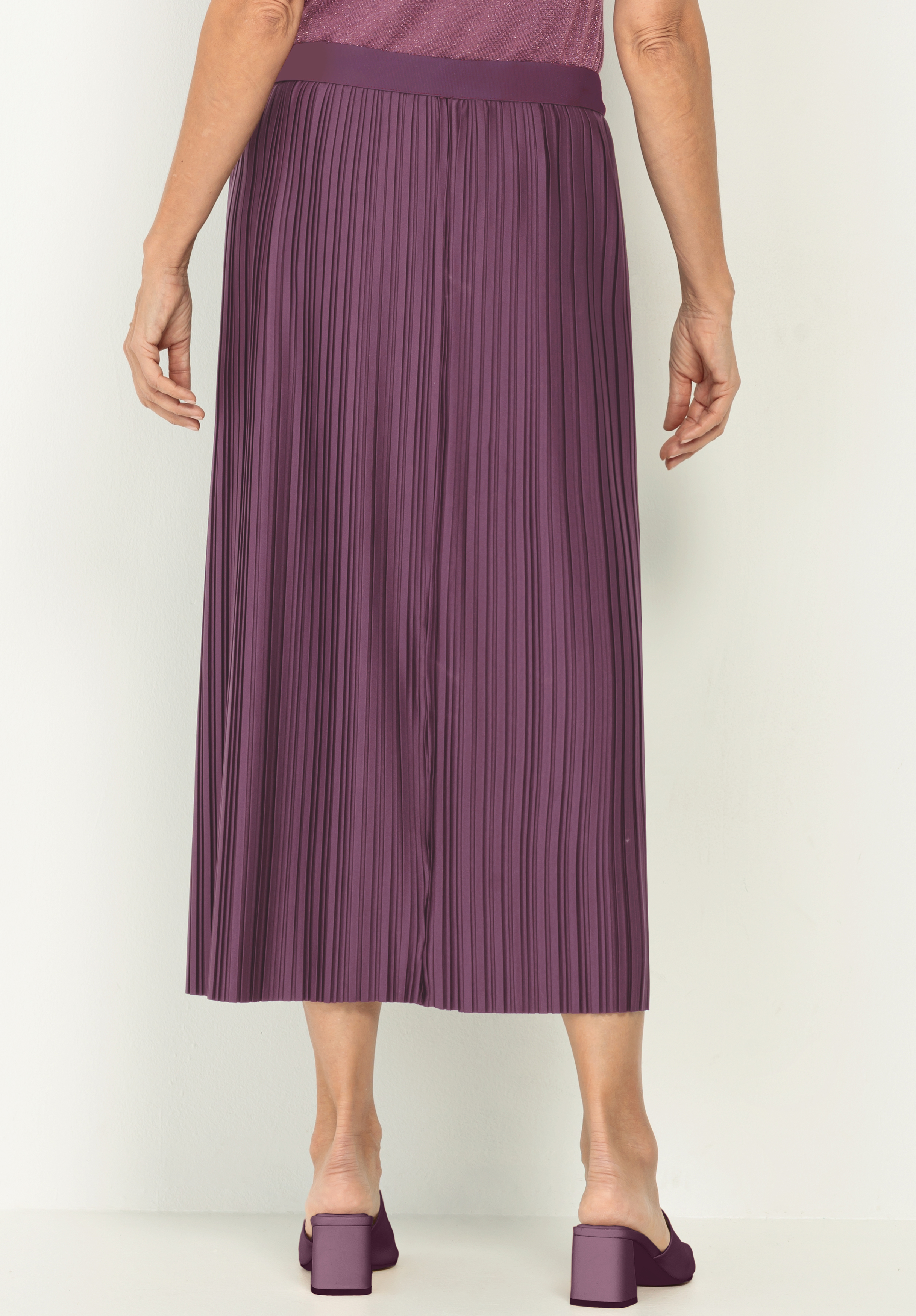 Eine Person steht mit dem Gesicht zur Wand und trägt einen lila Faltenrock und passende Schuhe mit Blockabsatz. Ihre Arme liegen entspannt an der Seite vor einem schlichten hellen Hintergrund.