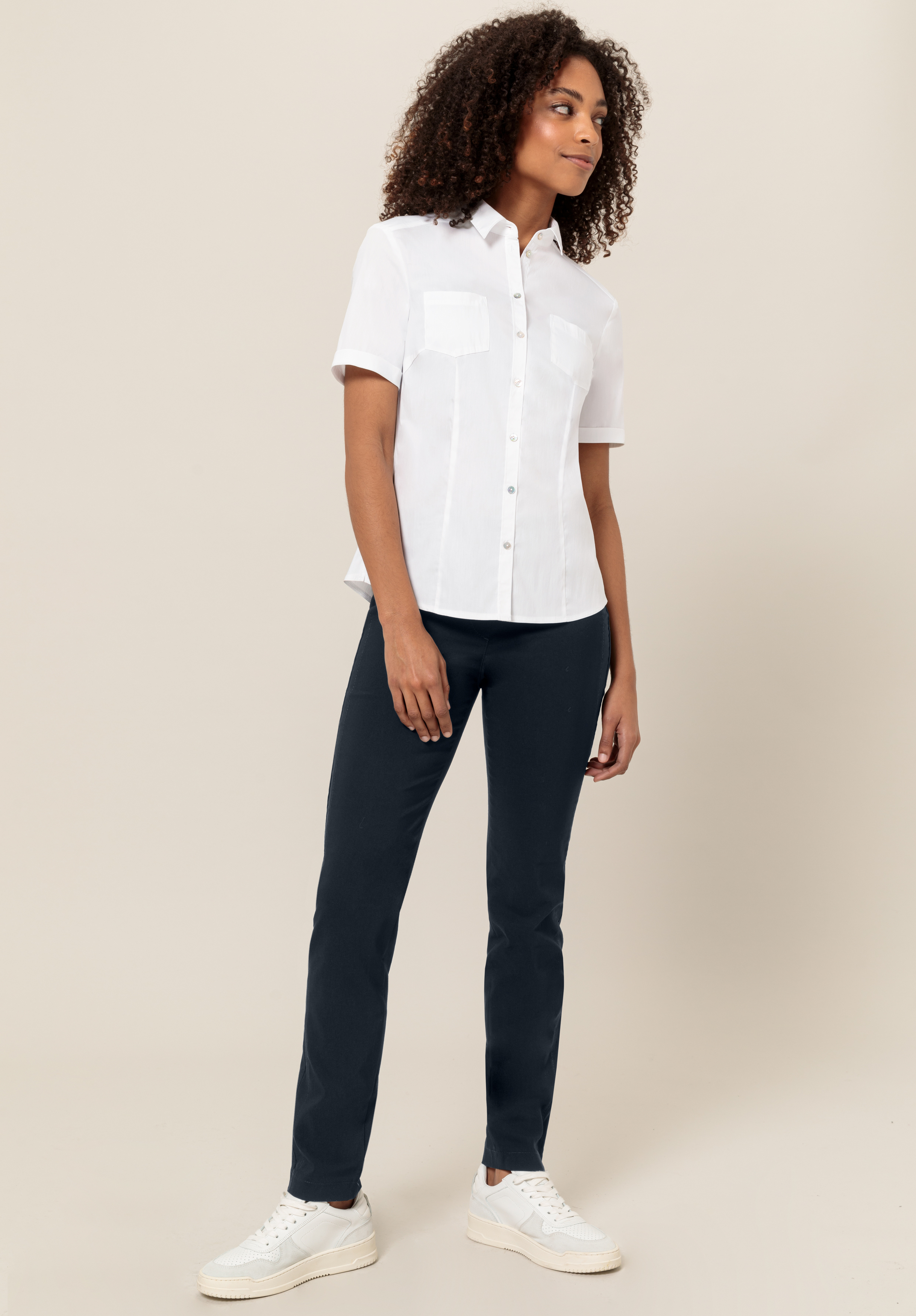 Eine Frau mit lockigem Haar, die ein kurzärmeliges weißes Button-up-Hemd, eine dunkelblaue Hose und weiße Turnschuhe trägt, steht vor einem schlichten beigen Hintergrund, blickt zur Seite und lächelt leicht.