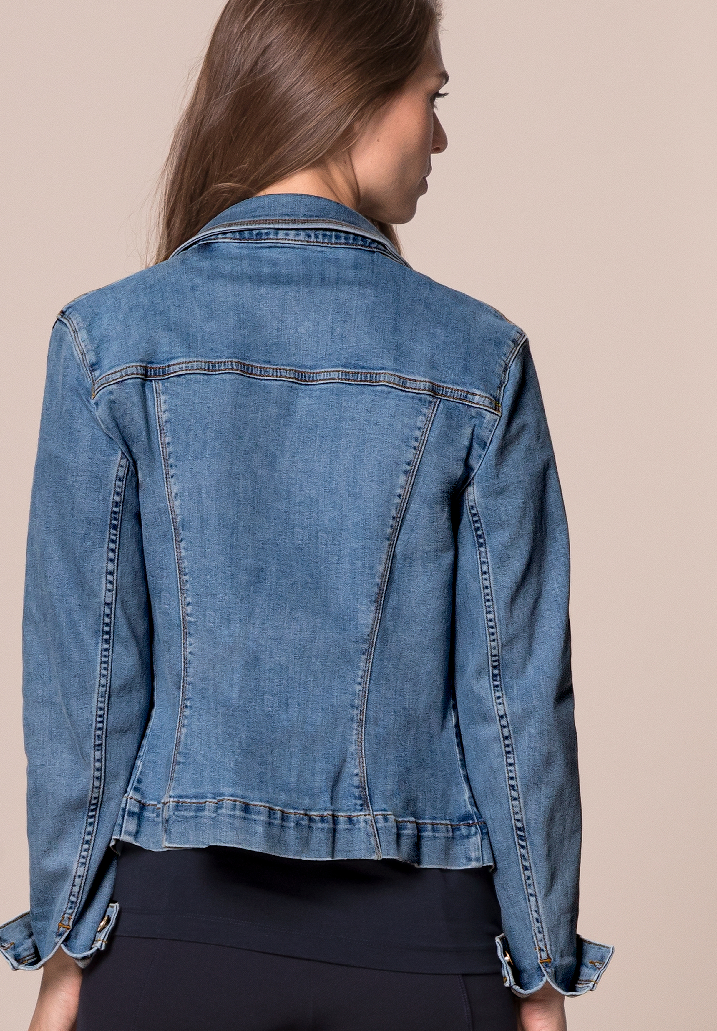 Eine Frau mit langen braunen Haaren wird von hinten gezeigt. Sie trägt eine blaue Jeansjacke und eine schwarze Hose vor einem schlichten hellbeigen Hintergrund.