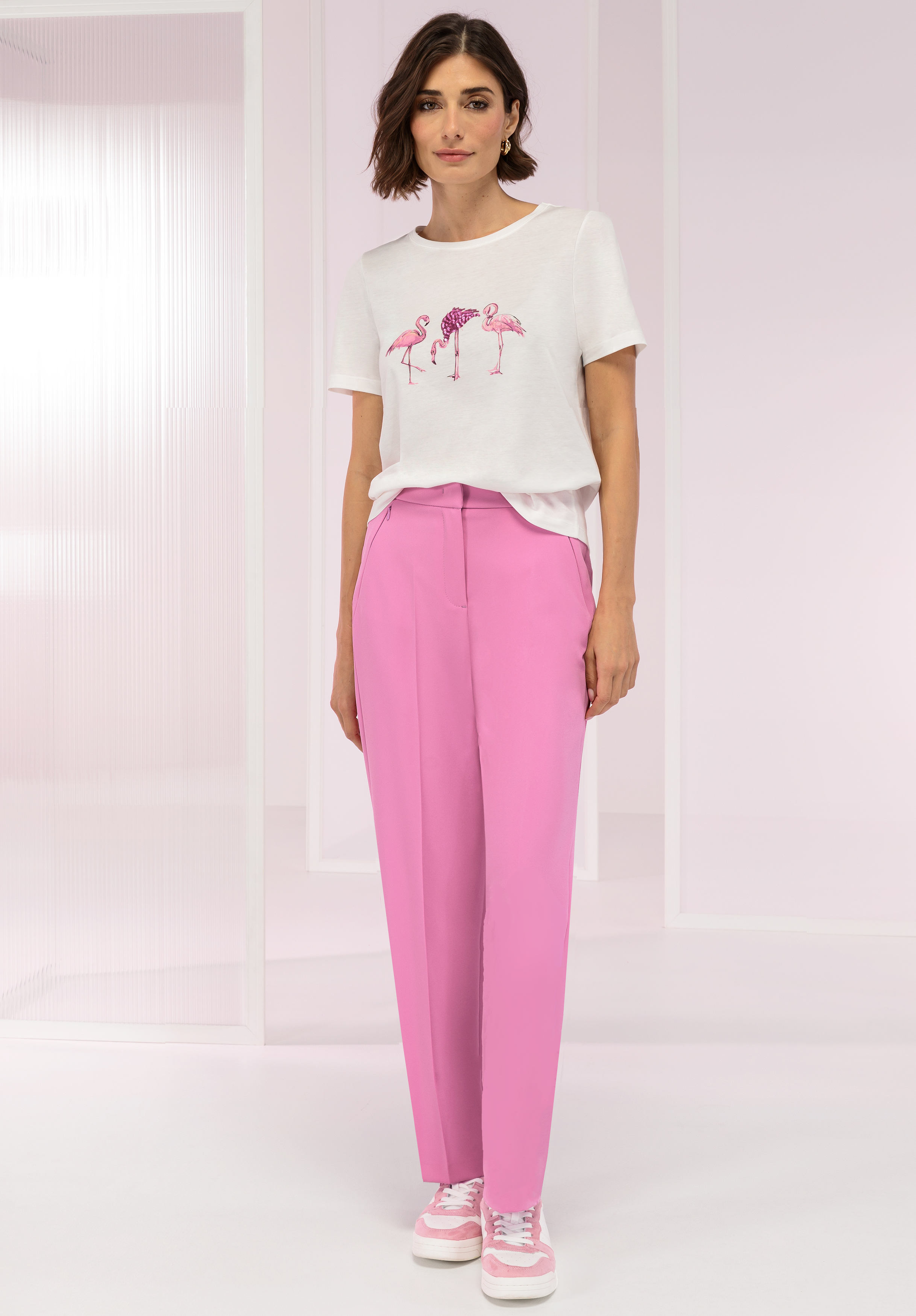 Eine Frau steht in einem hellrosa Raum. Sie trägt ein weißes T-Shirt mit rosa Flamingos, eine hoch taillierte rosa Hose und rosa-weiße Turnschuhe. Sie hat kurze braune Haare und eine entspannte Pose.