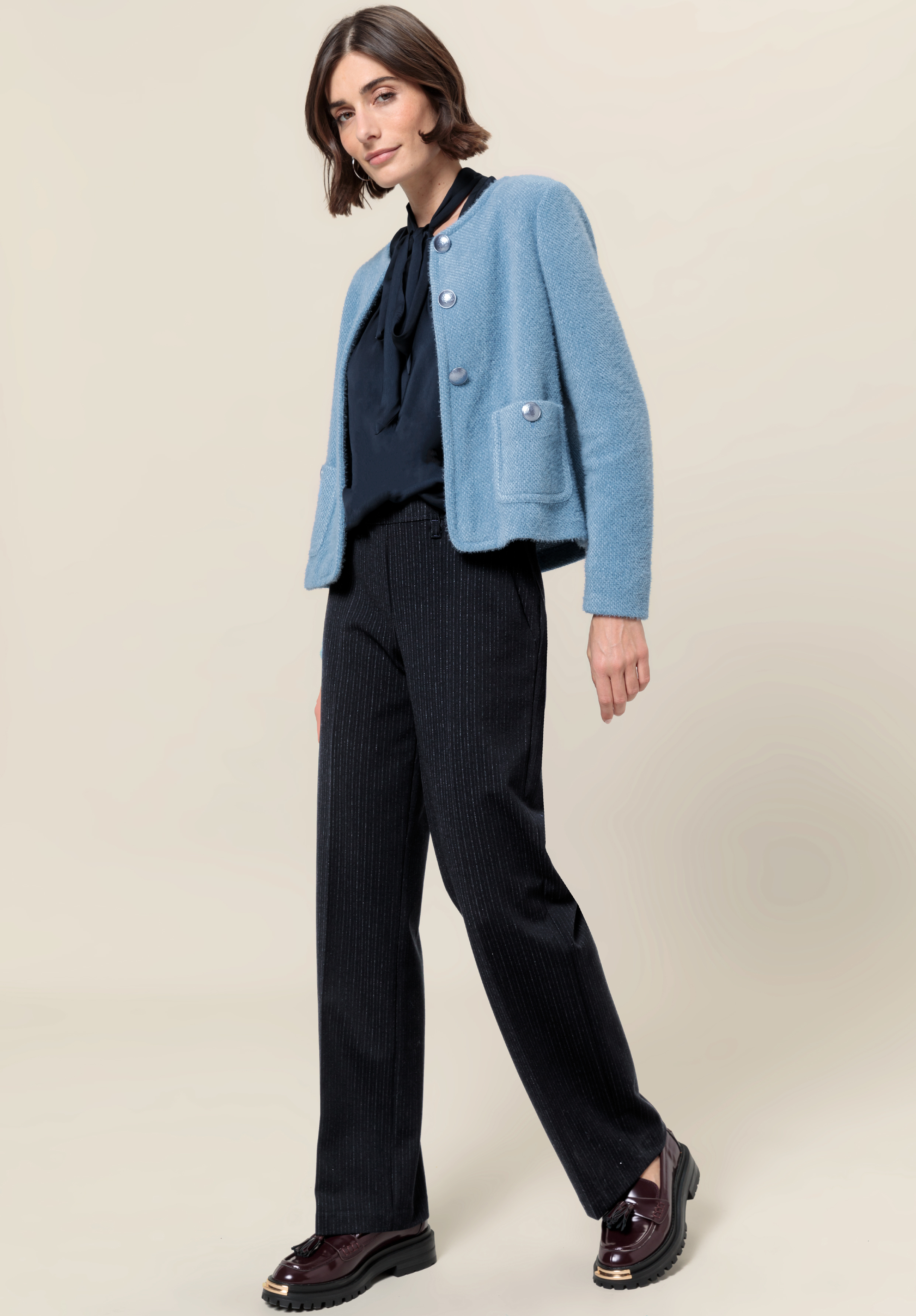Eine Person, die eine hellblaue Strickjacke über einer dunklen Bluse mit Schleife, eine schwarze Nadelstreifenhose und dunkle Slipper trägt, steht vor einem schlichten beigen Hintergrund und lächelt leicht.