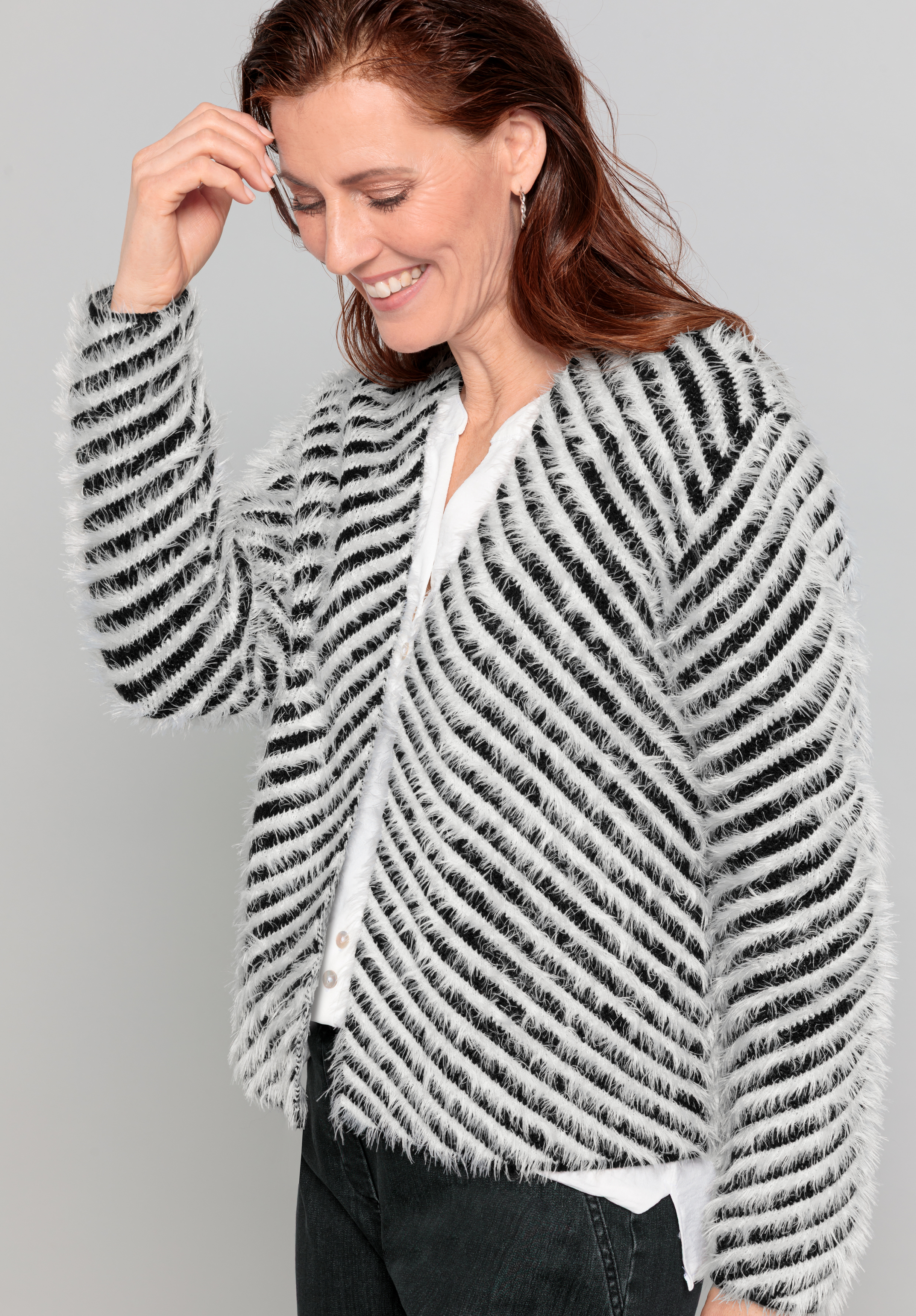 Eine Frau mit langen braunen Haaren lächelt, während sie nach unten blickt. Sie trägt einen flauschigen schwarz-weiß gestreiften Pullover über einem weißen Oberteil und einer schwarzen Hose und steht vor einem schlichten hellgrauen Hintergrund.