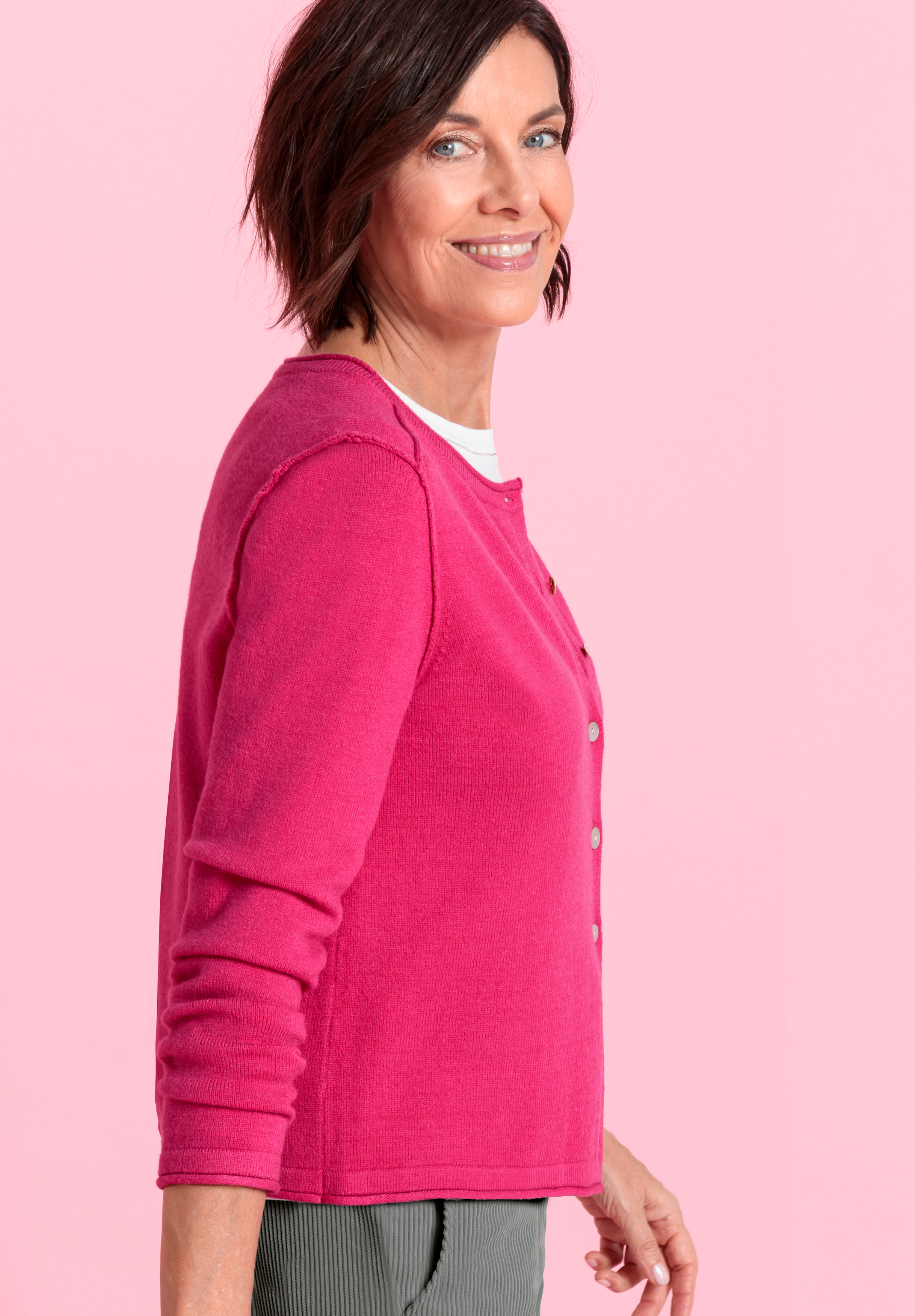 Eine Frau mit kurzen braunen Haaren, die eine leuchtend rosafarbene Strickjacke über einem weißen Oberteil und eine graue Hose trägt, steht seitlich und lächelt vor einem einfarbig rosa Hintergrund.