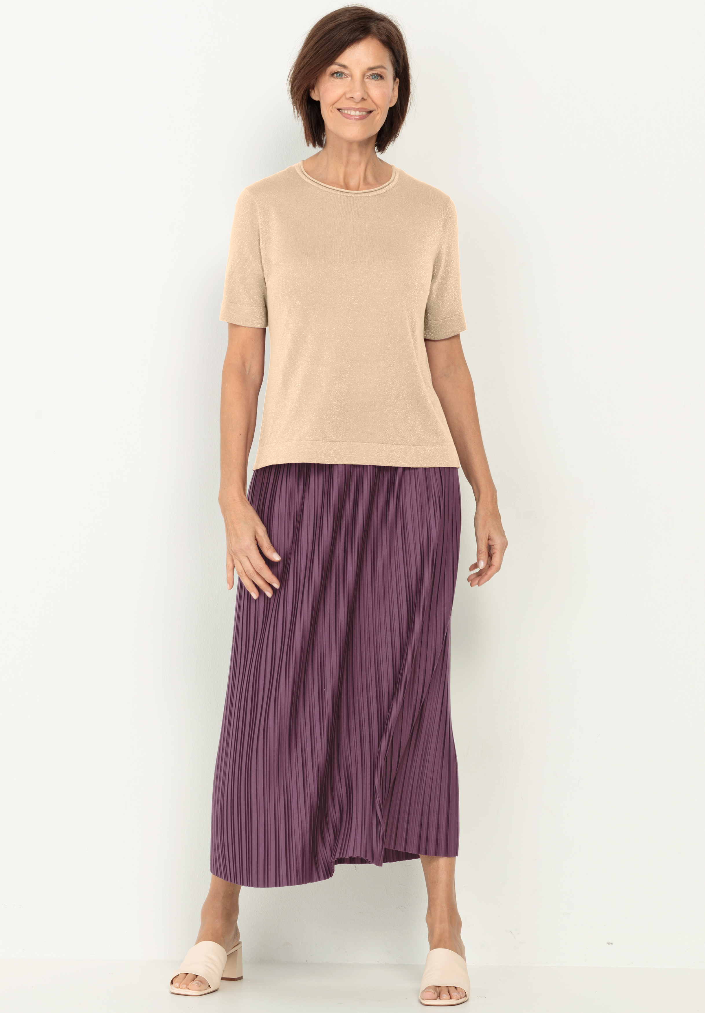 Eine Frau steht lächelnd vor einem weißen Hintergrund und trägt ein kurzärmeliges beiges Oberteil, einen langen lila Faltenrock und helle offene Schuhe.