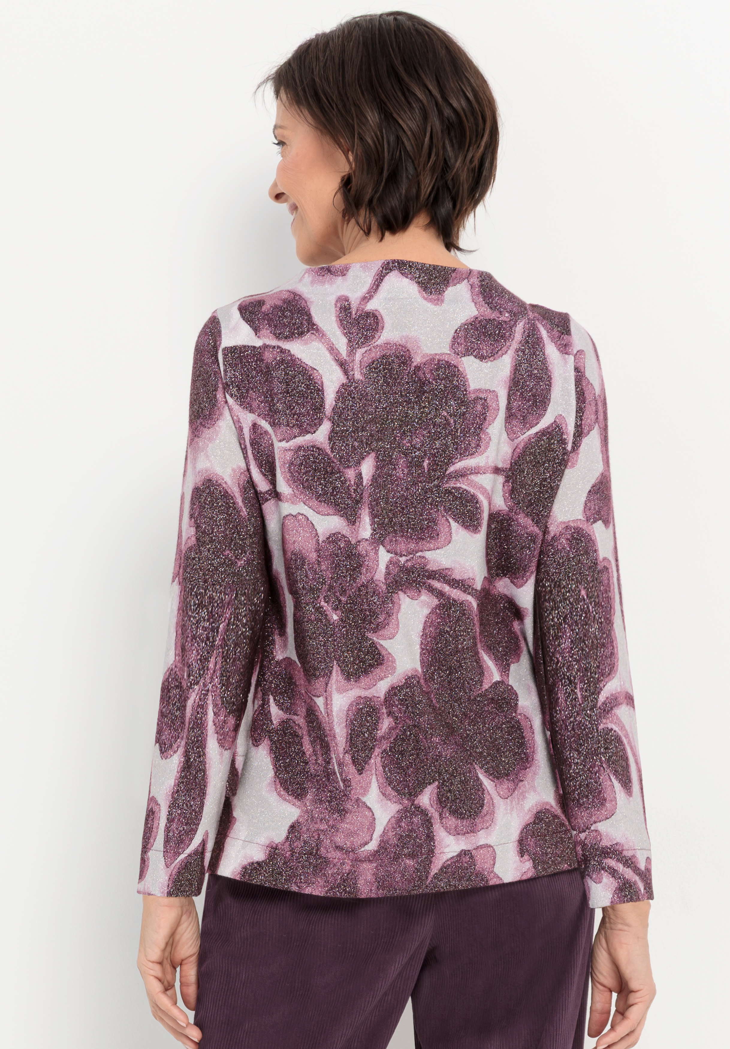 Eine Frau mit kurzen dunklen Haaren, von hinten gesehen, trägt ein langärmeliges Hemd mit einem großen lila Blumenmuster und eine dunkelviolette Hose. Sie lächelt und steht vor einem schlichten weißen Hintergrund.