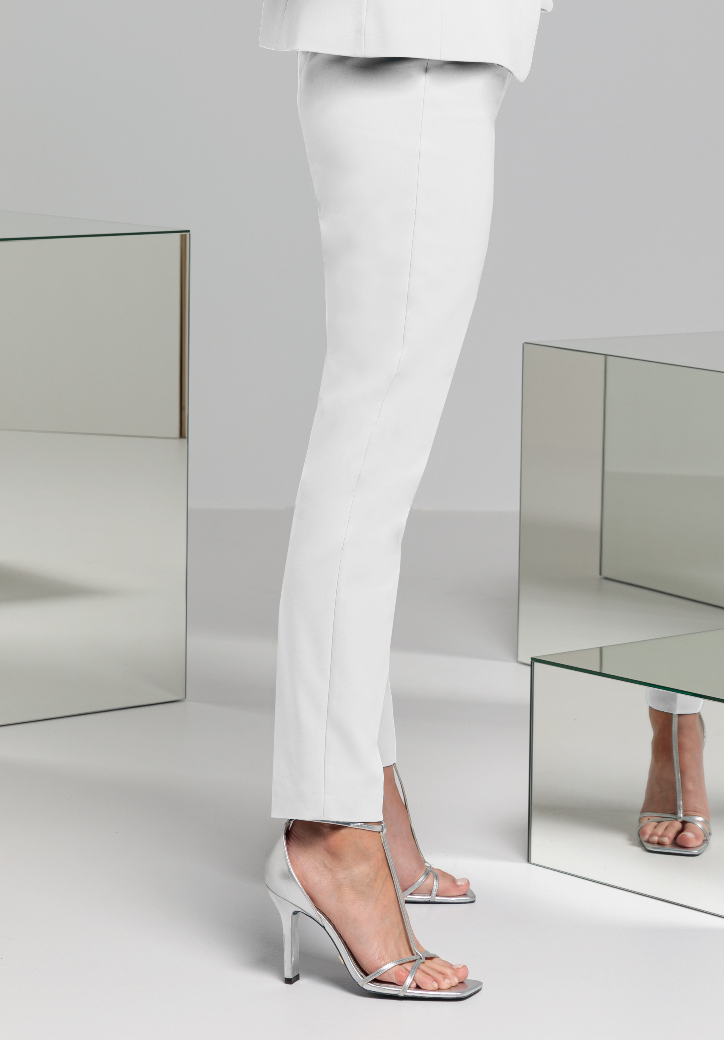 Eine Person in weißen Hosen und silbernen Sandalen mit hohen Absätzen steht neben verspiegelten Würfeln, auf denen ihr Spiegelbild sichtbar ist.