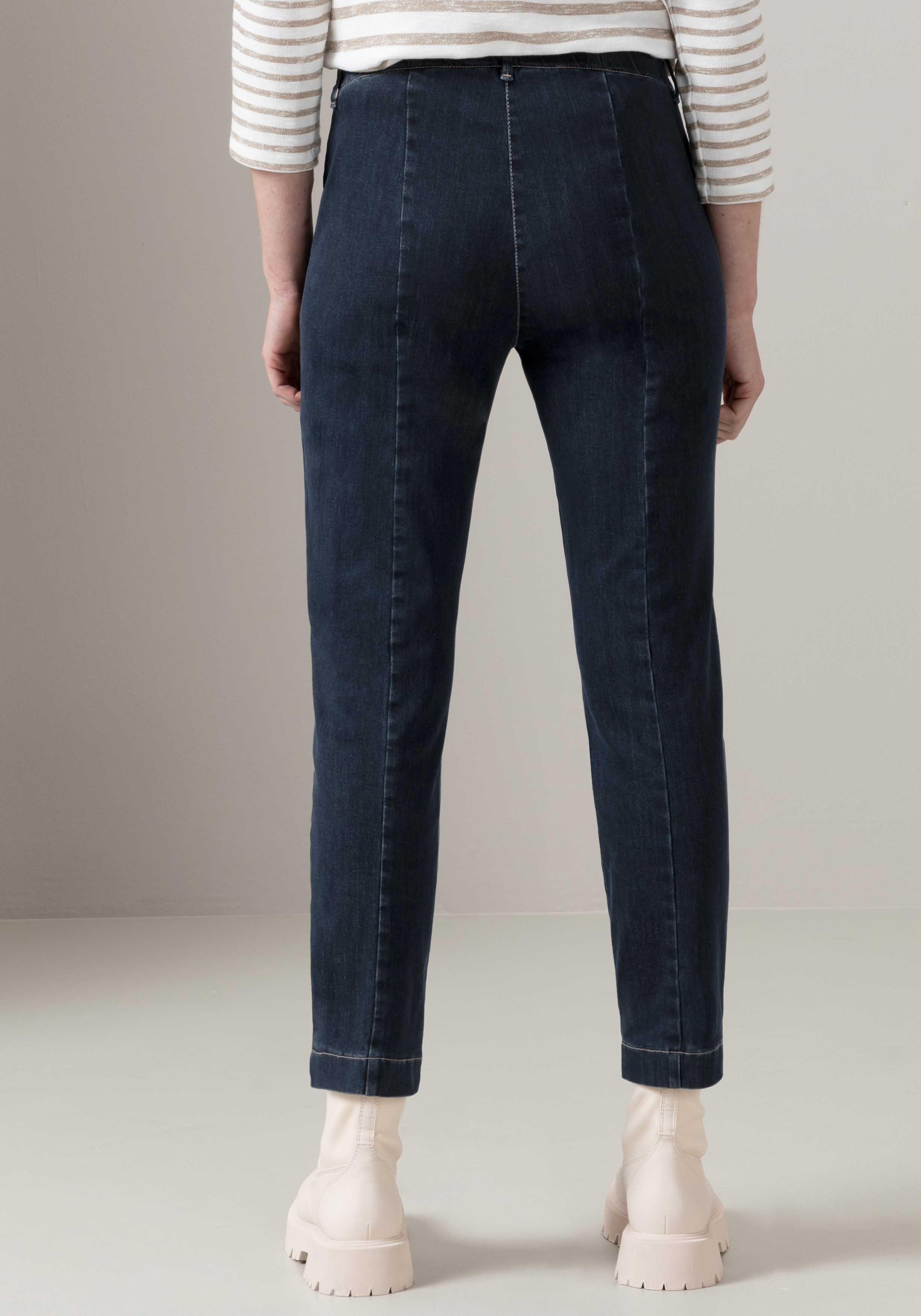 Eine Person, die dunkelblaue Jeans, ein weiß-beige gestreiftes Langarmshirt und hellbeige, klobige Stiefel trägt, steht mit dem Rücken zur Kamera vor einem schlichten, hellen Hintergrund.