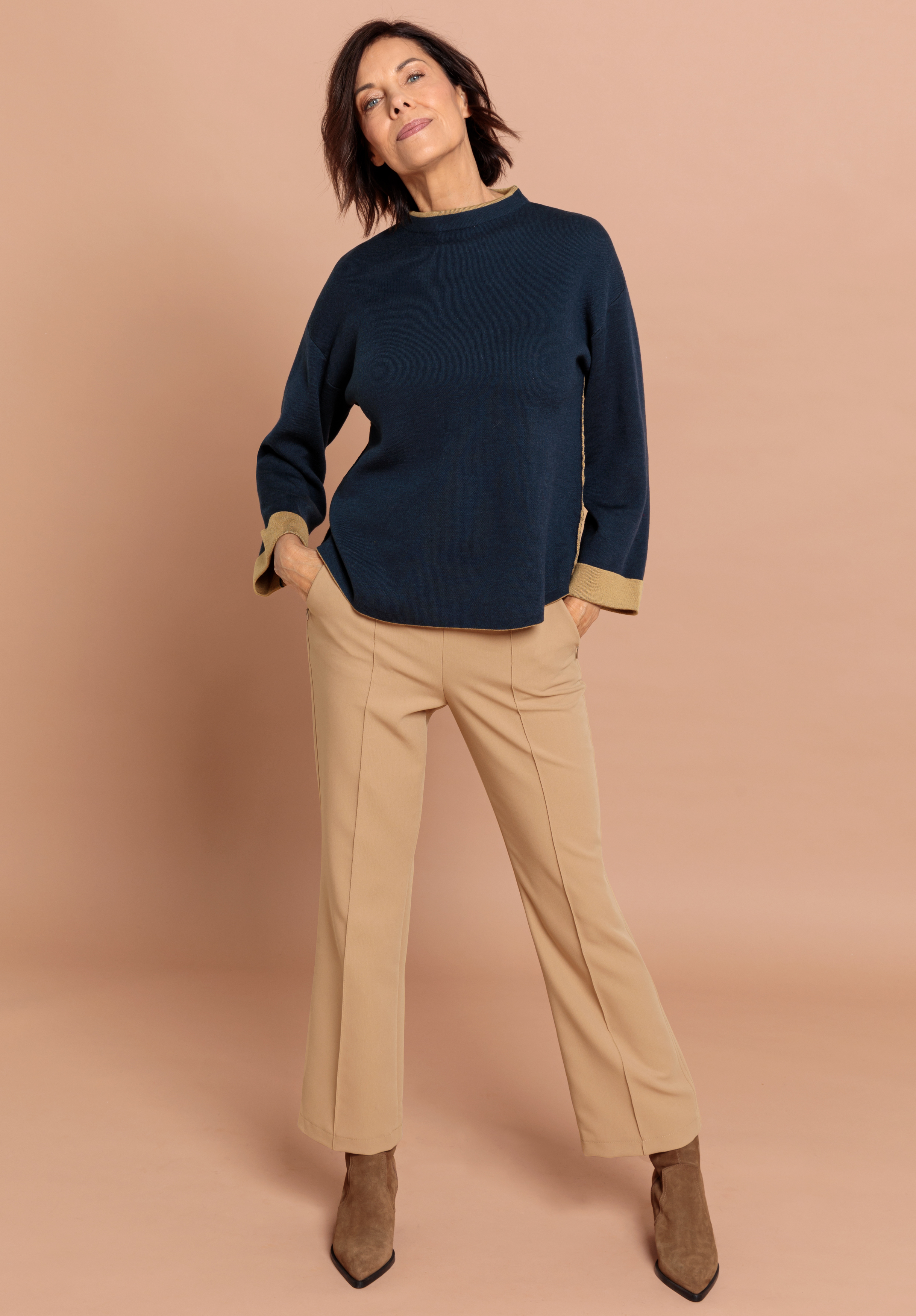 Eine Frau posiert selbstbewusst vor einem beigen Hintergrund. Sie trägt einen dunkelblauen, langärmeligen Pullover, eine beige Hose und braune, hochhackige Stiefel. Sie steht mit den Händen in den Taschen und lächelt leicht.