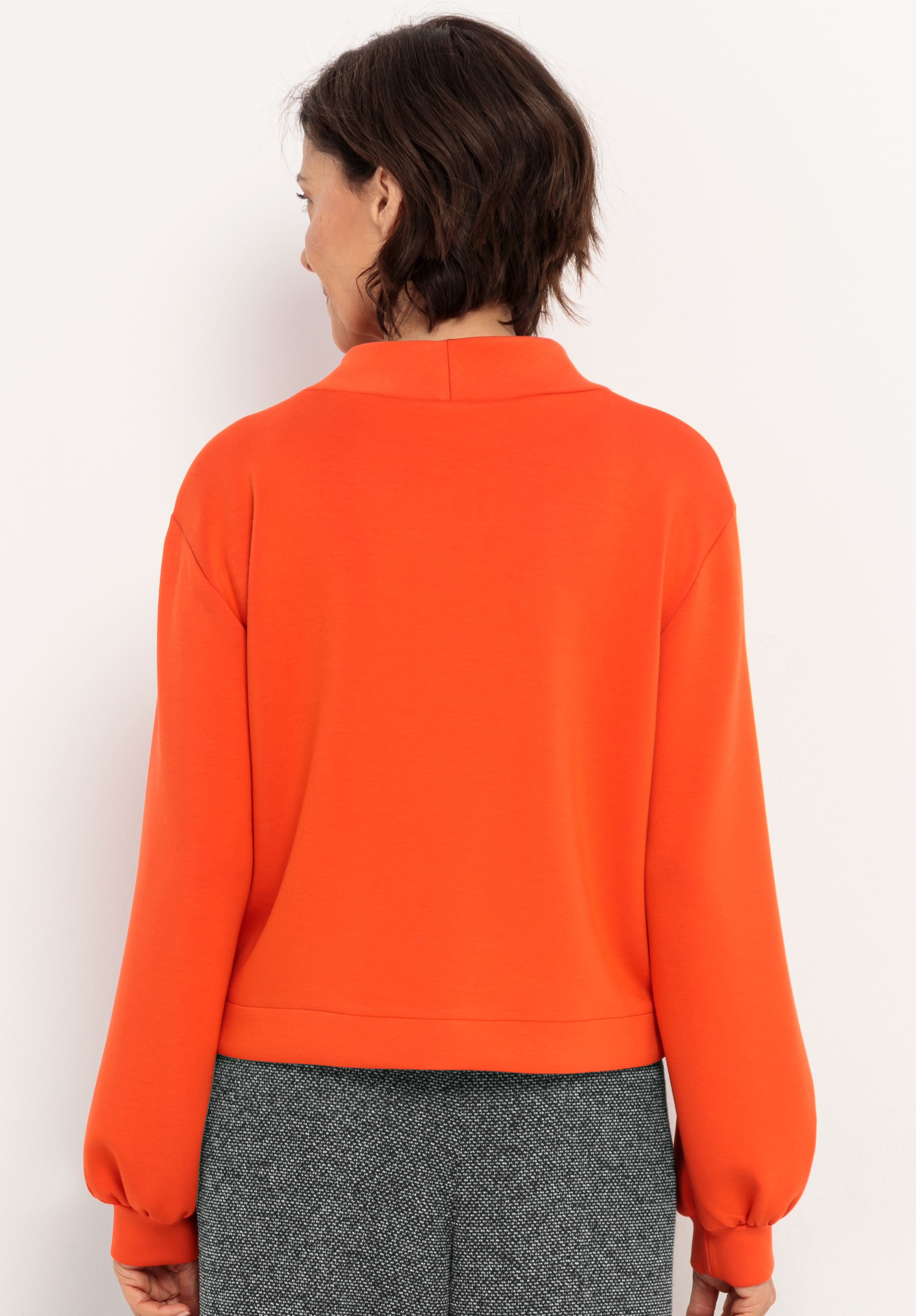 Eine Person mit kurzen braunen Haaren wird von hinten gezeigt, die ein orangefarbenes langärmeliges Oberteil und eine graue strukturierte Hose trägt und vor einem schlichten weißen Hintergrund steht.