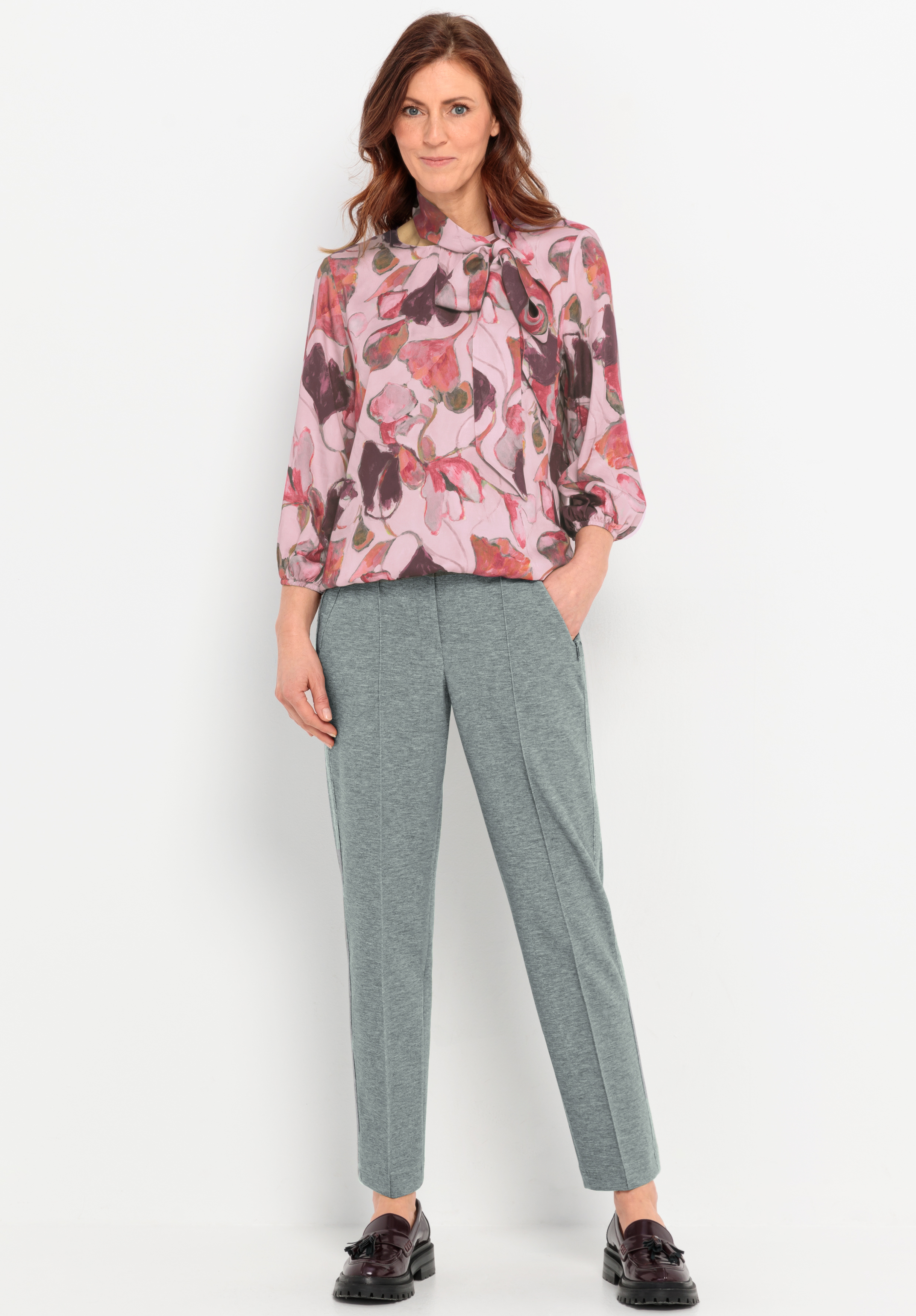 Eine Frau steht vor einem weißen Hintergrund und trägt eine rosa geblümte Bluse mit Ballonärmeln, eine graue maßgeschneiderte Hose und schwarze Halbschuhe. Sie hat eine Hand in der Tasche und lächelt sanft.