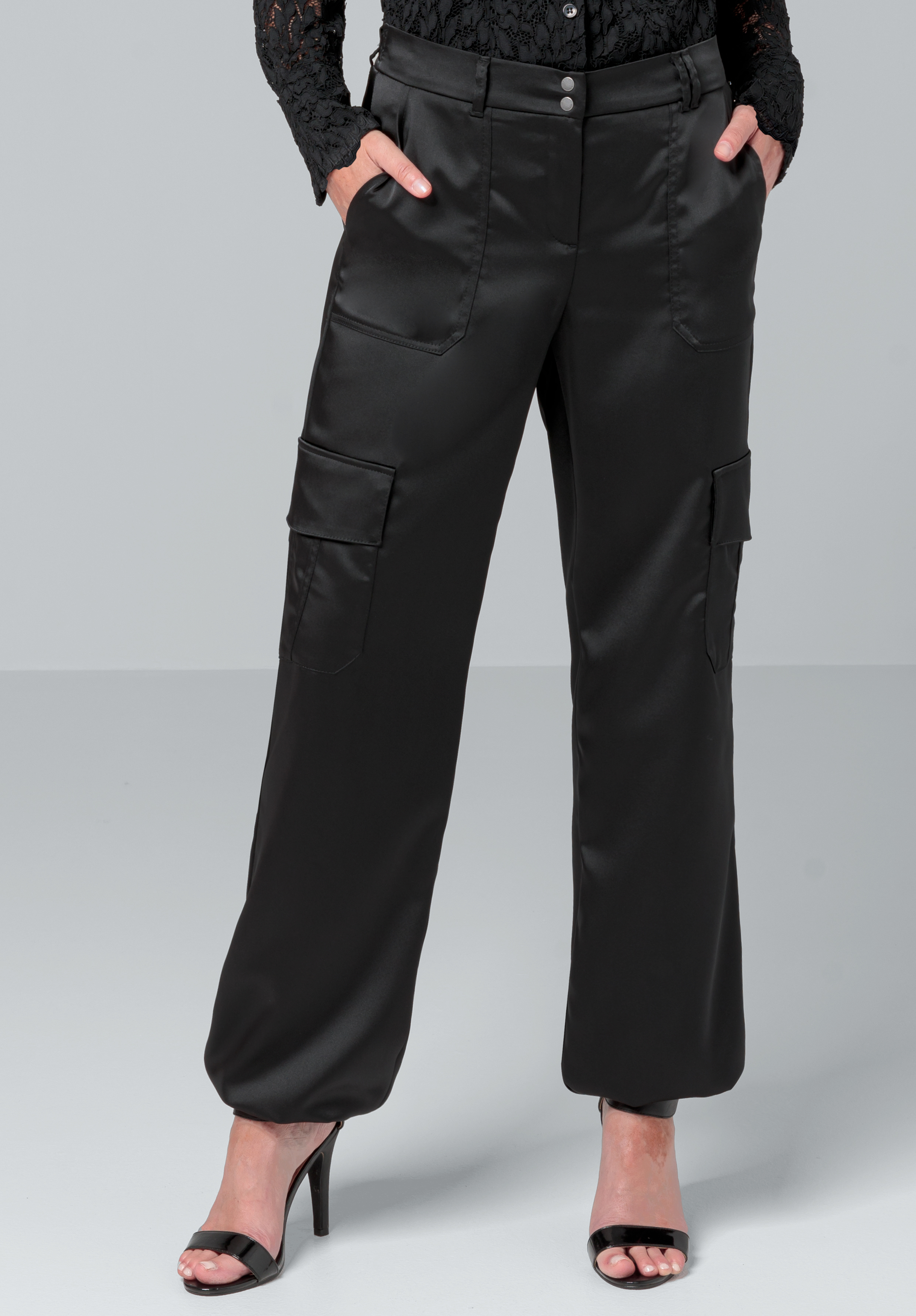 Eine Person in schwarzen Satin-Cargohosen mit großen Taschen, einem schwarzen Spitzentop und schwarzen offenen Pumps steht vor einem schlichten grauen Hintergrund. Nur der Unterkörper ist sichtbar.