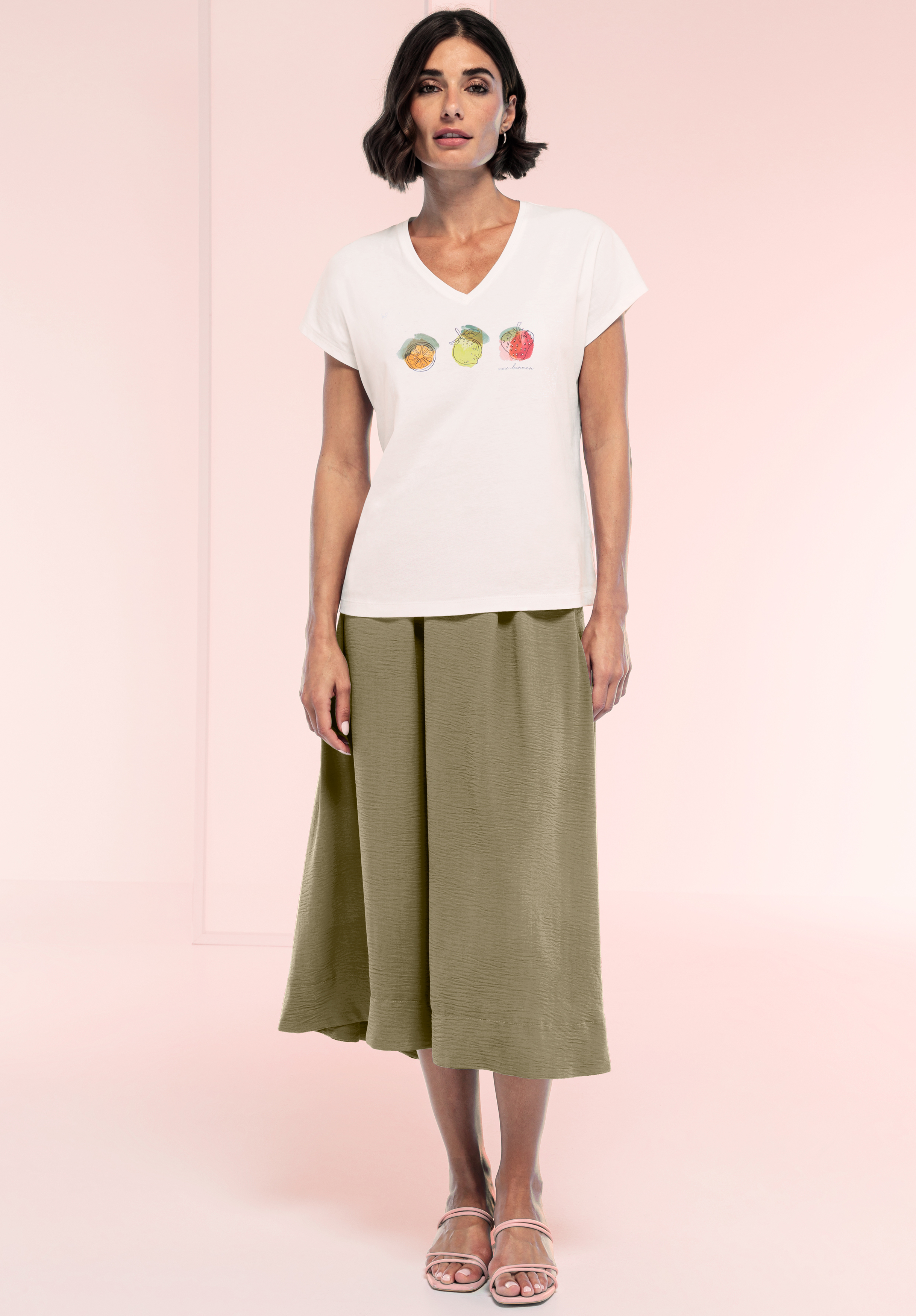 Eine Frau steht vor einem rosa Hintergrund. Sie trägt ein weißes T-Shirt mit drei Fruchtabbildungen, einen olivgrünen Midirock und helle Sandalen. Sie hat kurzes dunkles Haar und einen neutralen Gesichtsausdruck.