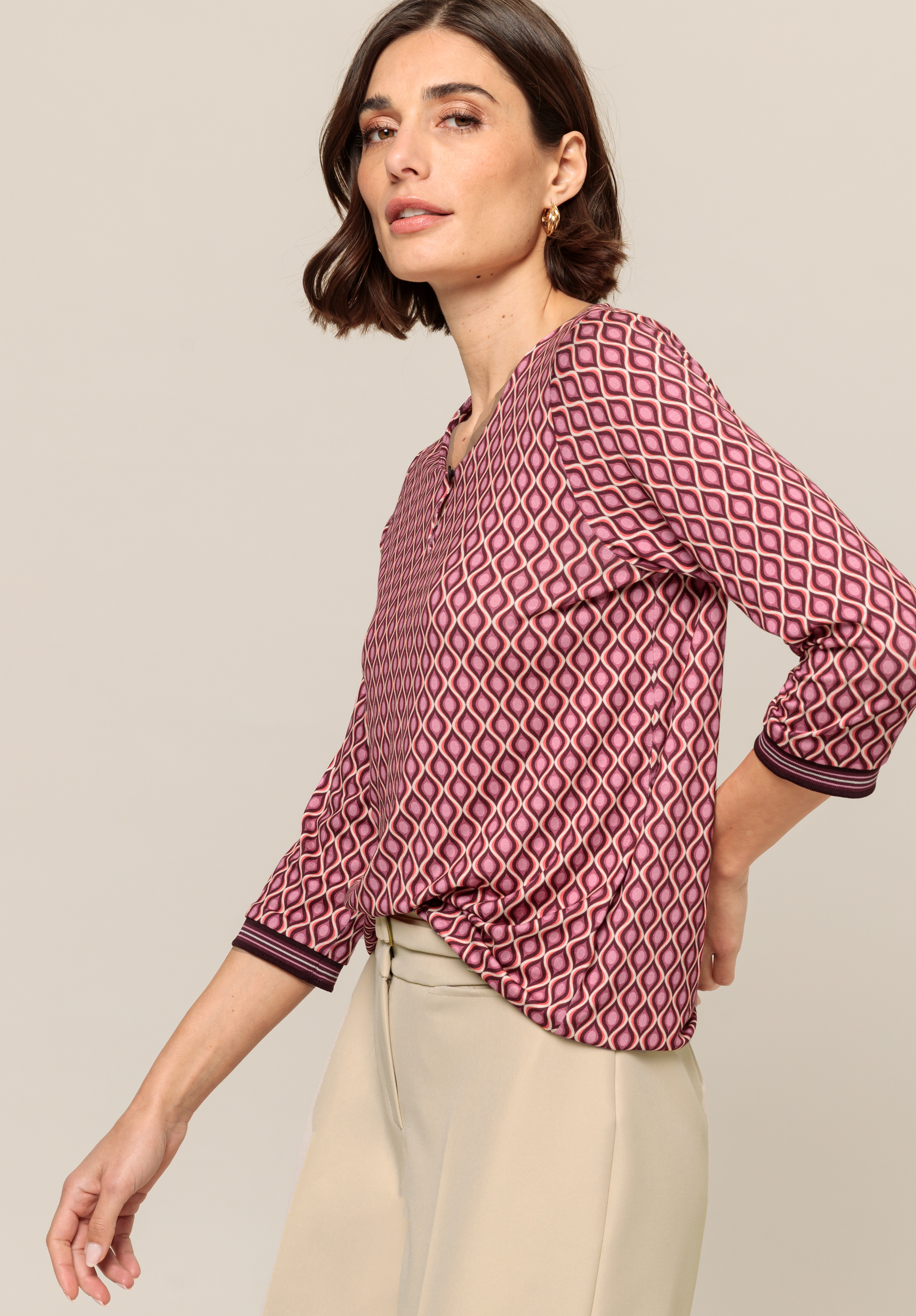 Eine Frau mit kurzen braunen Haaren trägt eine gemusterte Langarmbluse in Rosa- und Cremetönen, kombiniert mit einer hellbeigen Hose. Sie steht seitlich und blickt leicht über die Schulter.